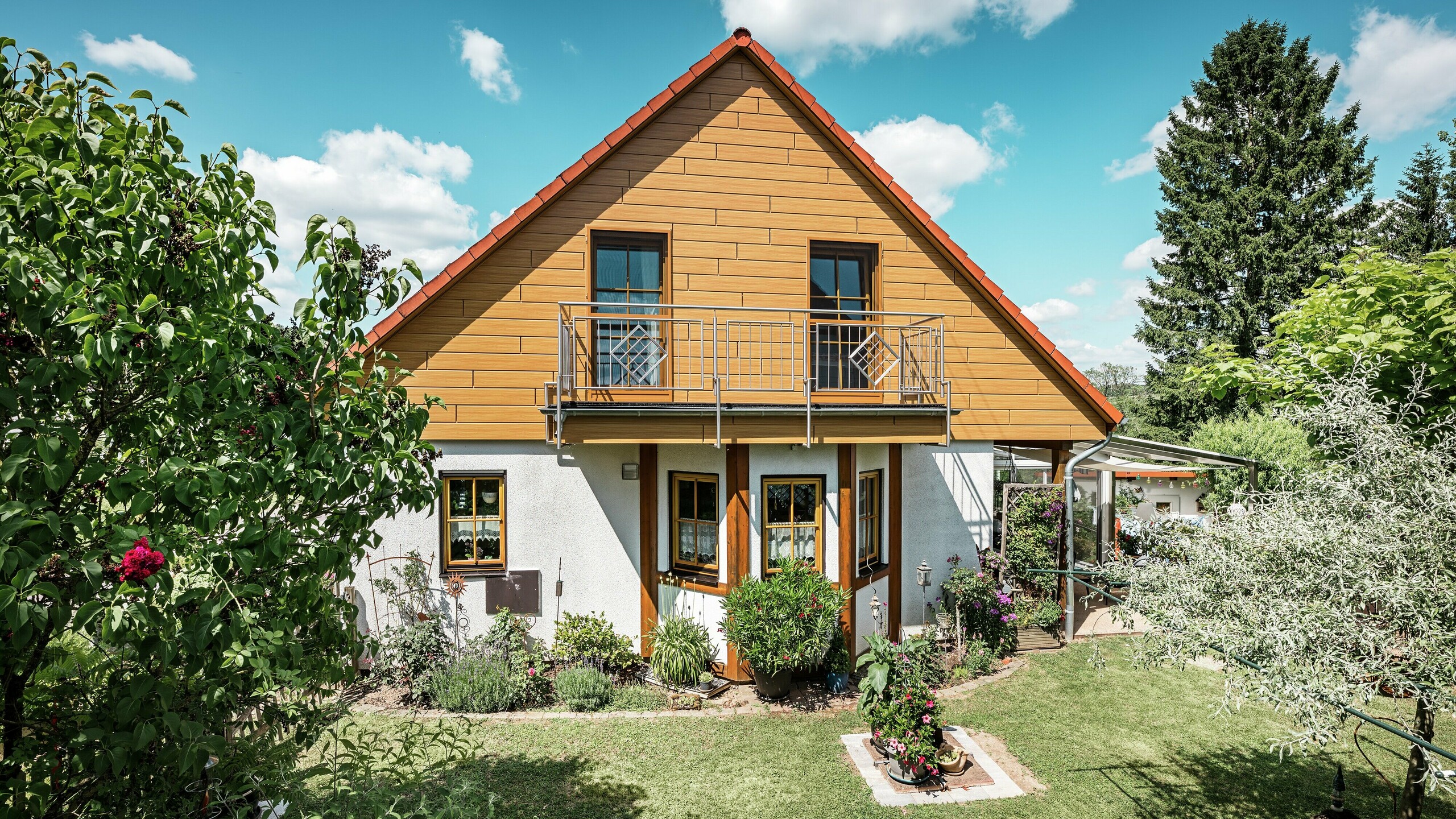 På dette billede stråler den renoverede gavl på et enfamiliehus i Blieskastel i en varm, natureg eg-tone takket være PREFA Siding. Den eksklusive aluminiumsfacade med realistisk trælook sætter et markant visuelt præg og harmonerer imponerende med det røde tegltag og den frodige have. Omgivet af blomster, buske og frodig grønne omgivelser fremstår huset indbydende og velplejet. PREFA-overfladen kombinerer et naturligt udseende med funktionelle fordele som høj vejrbestandighed, stabilitet og næsten vedligeholdelsesfri holdbarhed – et æstetisk højdepunkt med bæredygtig merværdi.