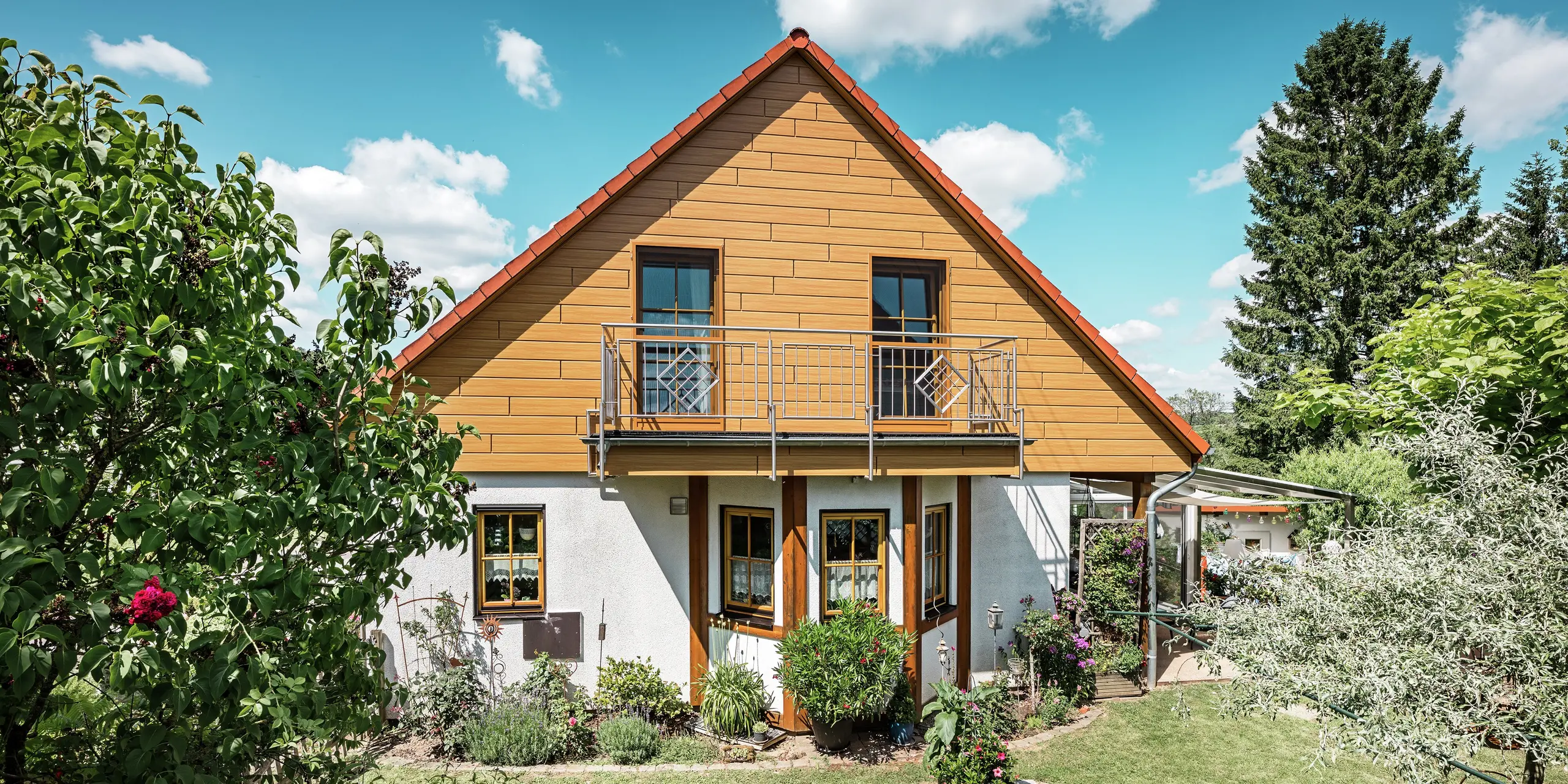 På dette billede stråler den renoverede gavl på et enfamiliehus i Blieskastel i en varm, natureg eg-tone takket være PREFA Siding. Den eksklusive aluminiumsfacade med realistisk trælook sætter et markant visuelt præg og harmonerer imponerende med det røde tegltag og den frodige have. Omgivet af blomster, buske og frodig grønne omgivelser fremstår huset indbydende og velplejet. PREFA-overfladen kombinerer et naturligt udseende med funktionelle fordele som høj vejrbestandighed, stabilitet og næsten vedligeholdelsesfri holdbarhed – et æstetisk højdepunkt med bæredygtig merværdi.