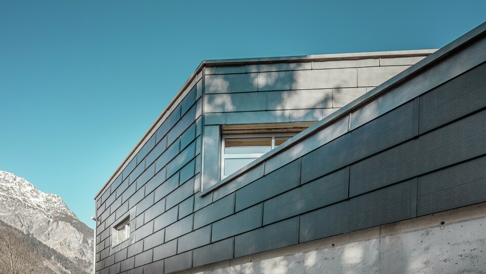 Die Fassade des Bürogebäudes der RÜMA Blechwerkstatt in Vorarlberg mit innovativen PREFA Solardachpaneelen und Sichtbeton, umgeben von Bergen unter klarem blauem Himmel, mit Baumschatten an der Wand.