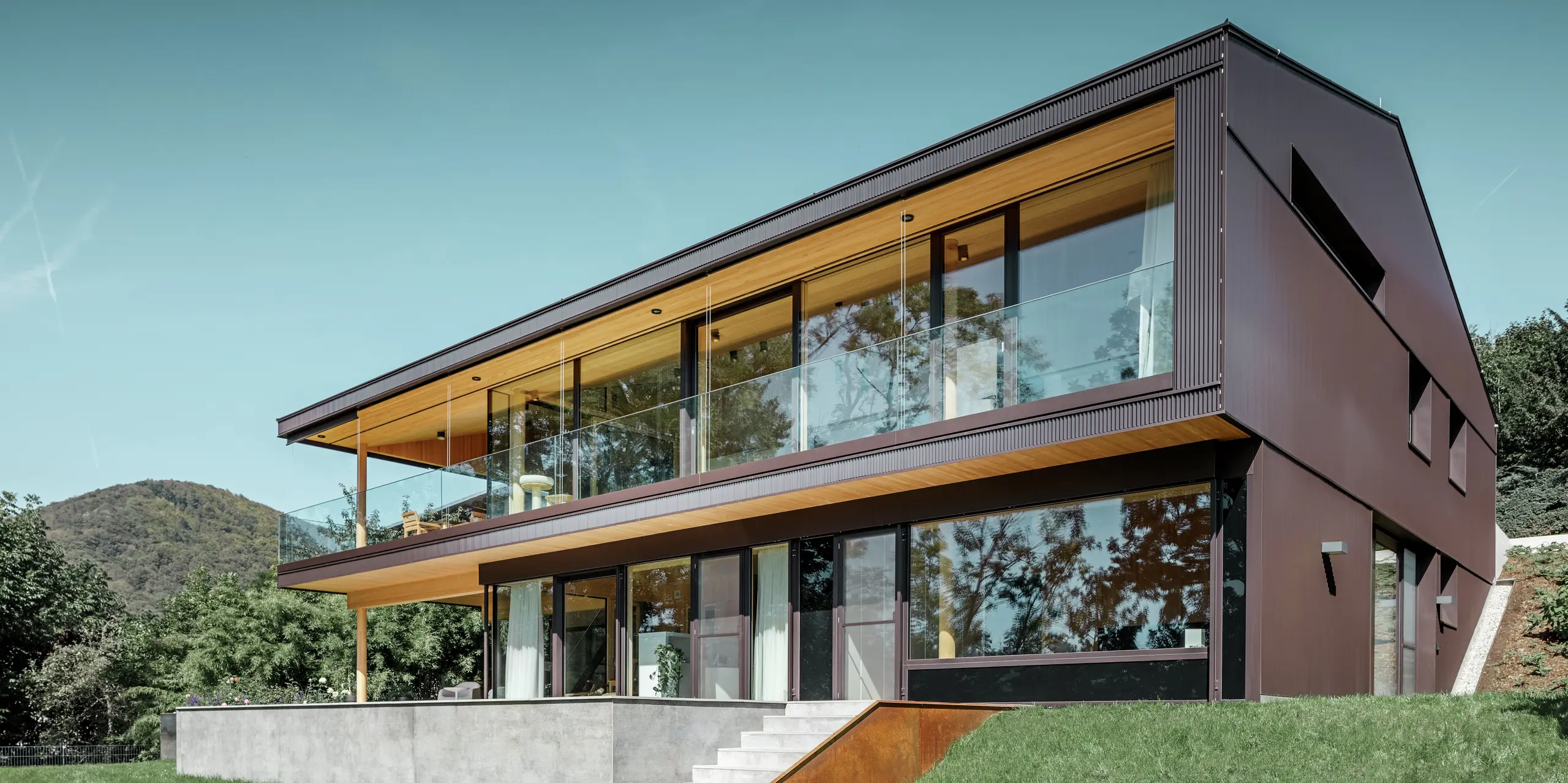 Dette moderne enfamilieshus befinder sig midt i naturen. Bygningens facade blev beklædt med PREFA-takker i farven nøddebrun. Beklædningen passer derved både til huset og til det omliggende landskab.
