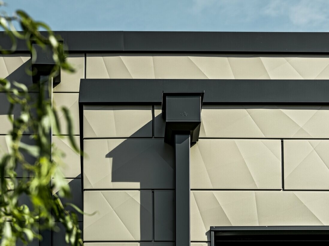 Moderne parcelhus med fladt tag og smalle vinduer. Facaden er beklædt med PREFA Siding.X-facade med rynket udseende i farven bronze. Forbindelserne blev lavet i antracit. PREFA-kvadratrør med tilhørende vandopsamlingsbeholder i antracit bruges til tagafvanding.