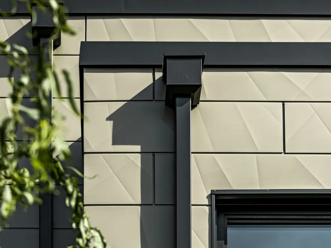 Moderne parcelhus med fladt tag og smalle vinduer. Facaden er beklædt med PREFA Siding.X-facade med rynket udseende i farven bronze. Forbindelserne blev lavet i antracit. PREFA-kvadratrør med tilhørende vandopsamlingsbeholder i antracit bruges til tagafvanding.