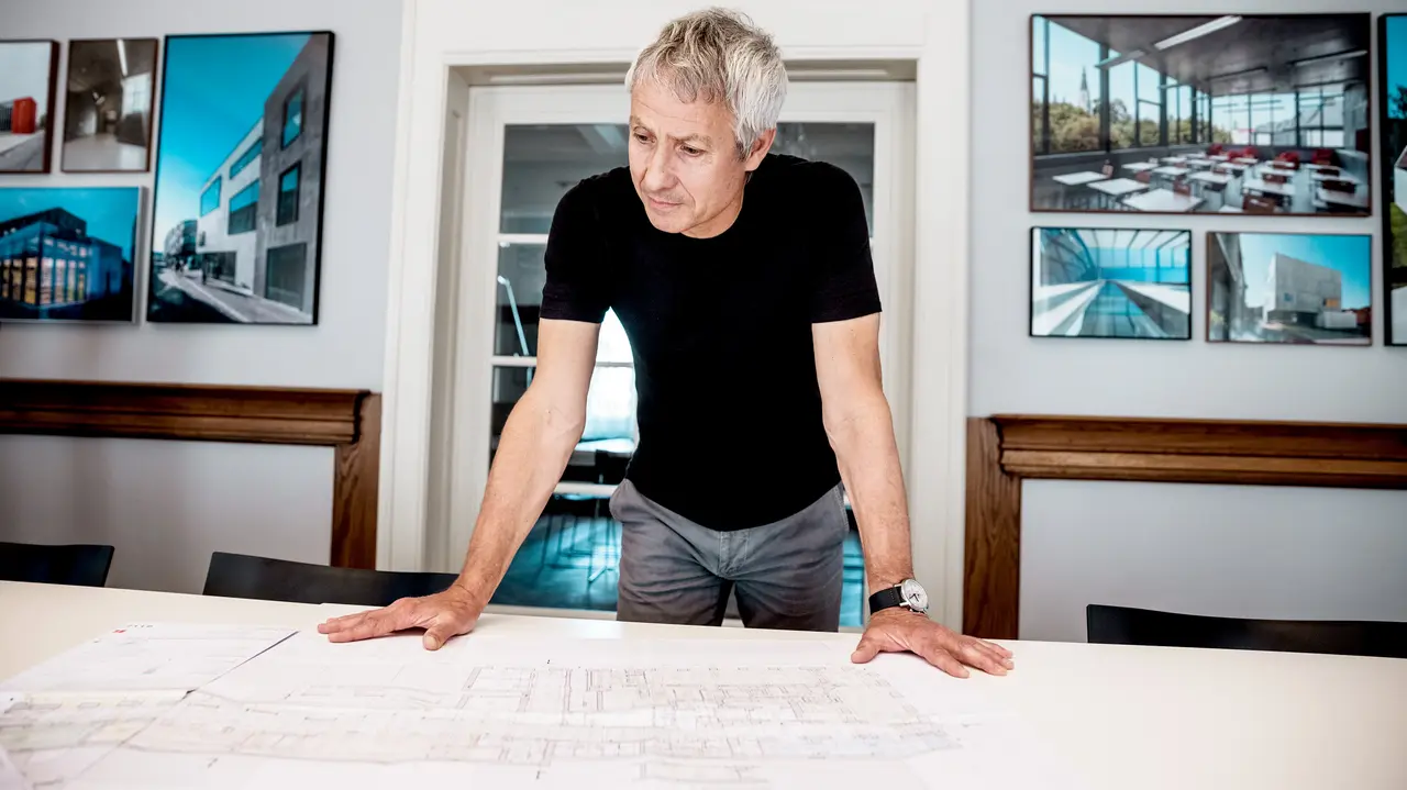 Arkitekten arbejder på detaljerede byggeplaner i et moderne kontor, omgivet af imponerende fotos af PREFA-arkitekturprojekter.