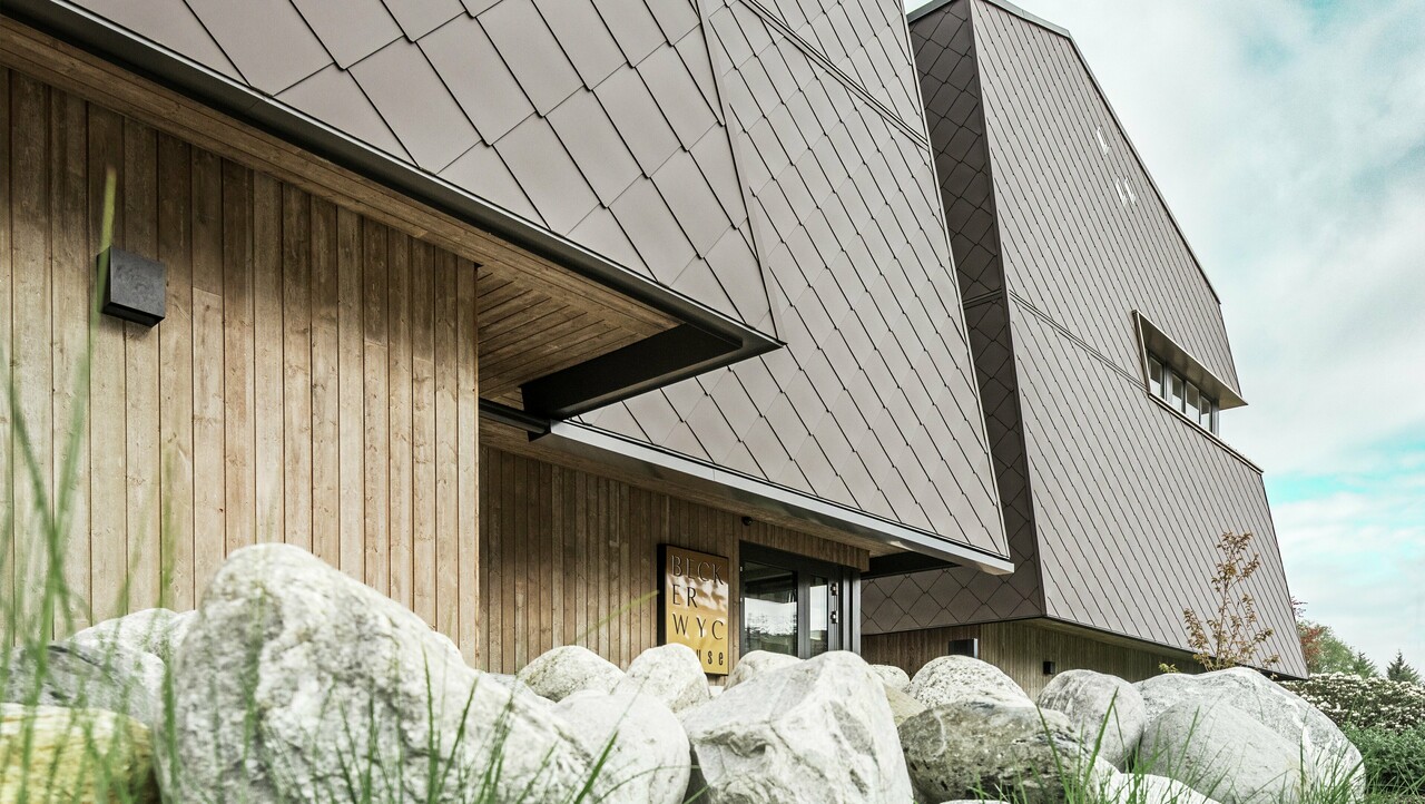 Sidebillede af et hotel, stueetagens facade er beklædt lodret med mørkfarvet træ. Over dette er der et facadefremspring, og herfra er facaden beklædt med aluminiumspaneler i valnøddebrun.