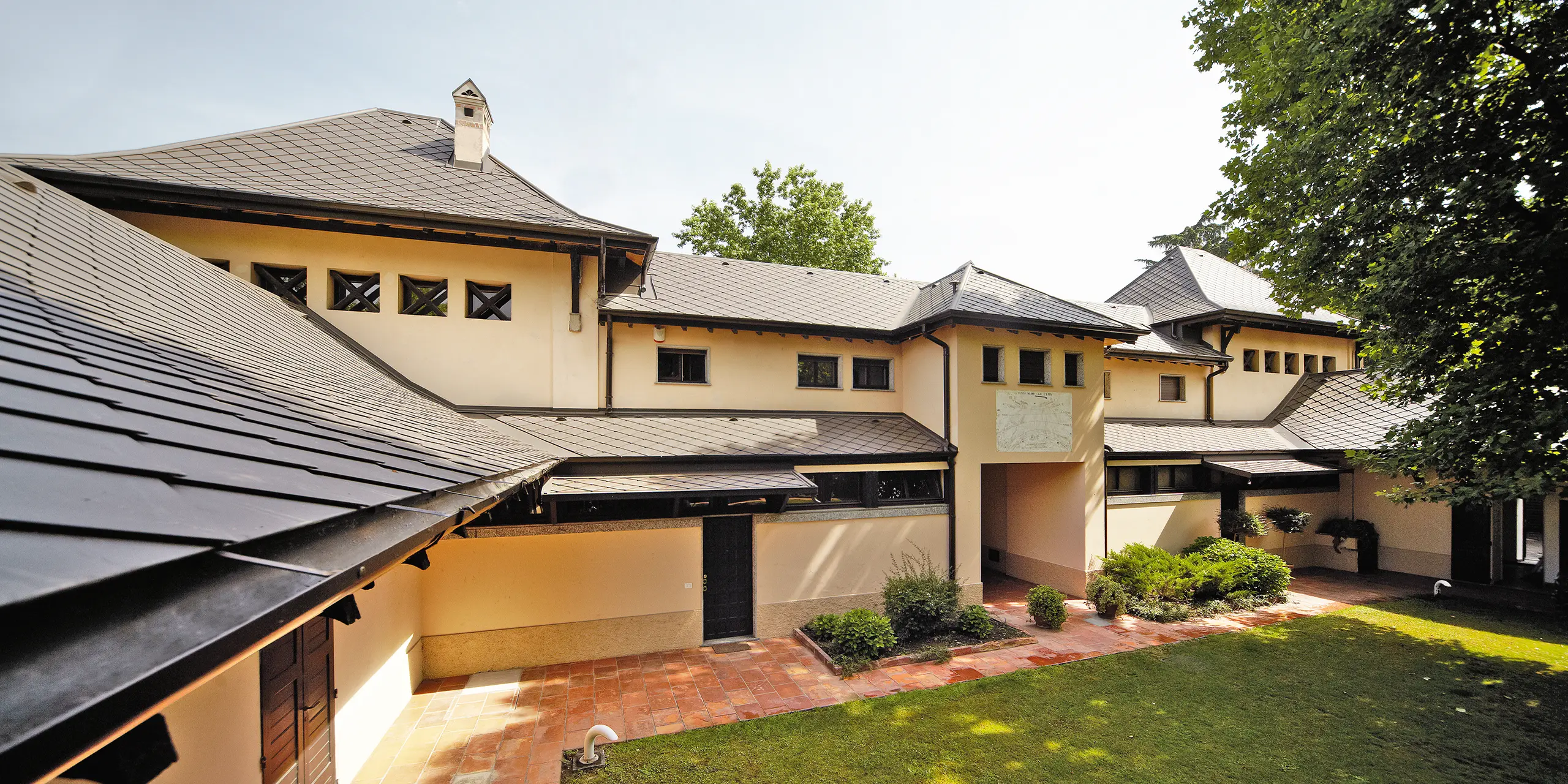 Italiensk villa med rombetag i P.10 brun fra PREFA med beige facadefarve og skyggefuld baghave