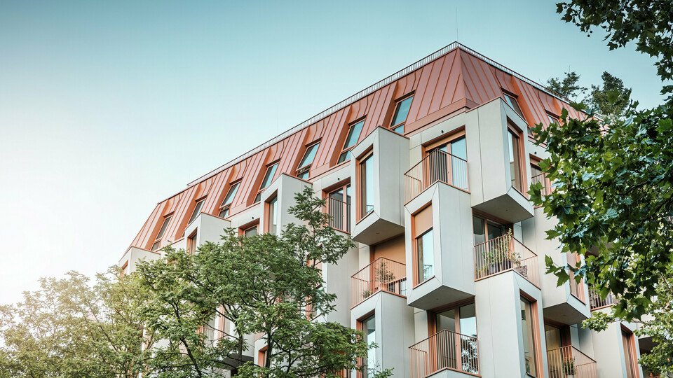 Moderne Wohnarchitektur im Projekt Van B in München mit einem innovativen Erker und einem markanten PREFALZ-Aluminiumdach in der Sonderfarbe IGP Orange HDP von PREFA. Das hochwertige Dachmaterial überzeugt durch seine außergewöhnliche Langlebigkeit, hohe Widerstandsfähigkeit und Farbstabilität. In Kombination mit der kubischen Fassadenform und den großflächigen Glasfronten entsteht ein harmonisches Gesamtbild, das Funktionalität, Design und Nachhaltigkeit auf höchstem Niveau vereint – ideal für urbanes Wohnen.