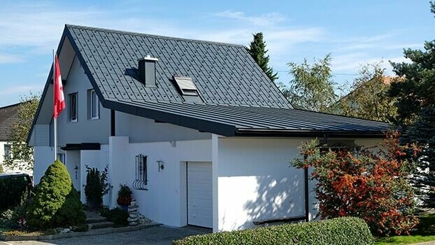 Renoveret hus med saddeltag og tilhørende garage. Taget blev dækket med PREFA tagplader og garagen med prefalz i antracit. Foran huset ses en flagstang med det Schweiziske flag.