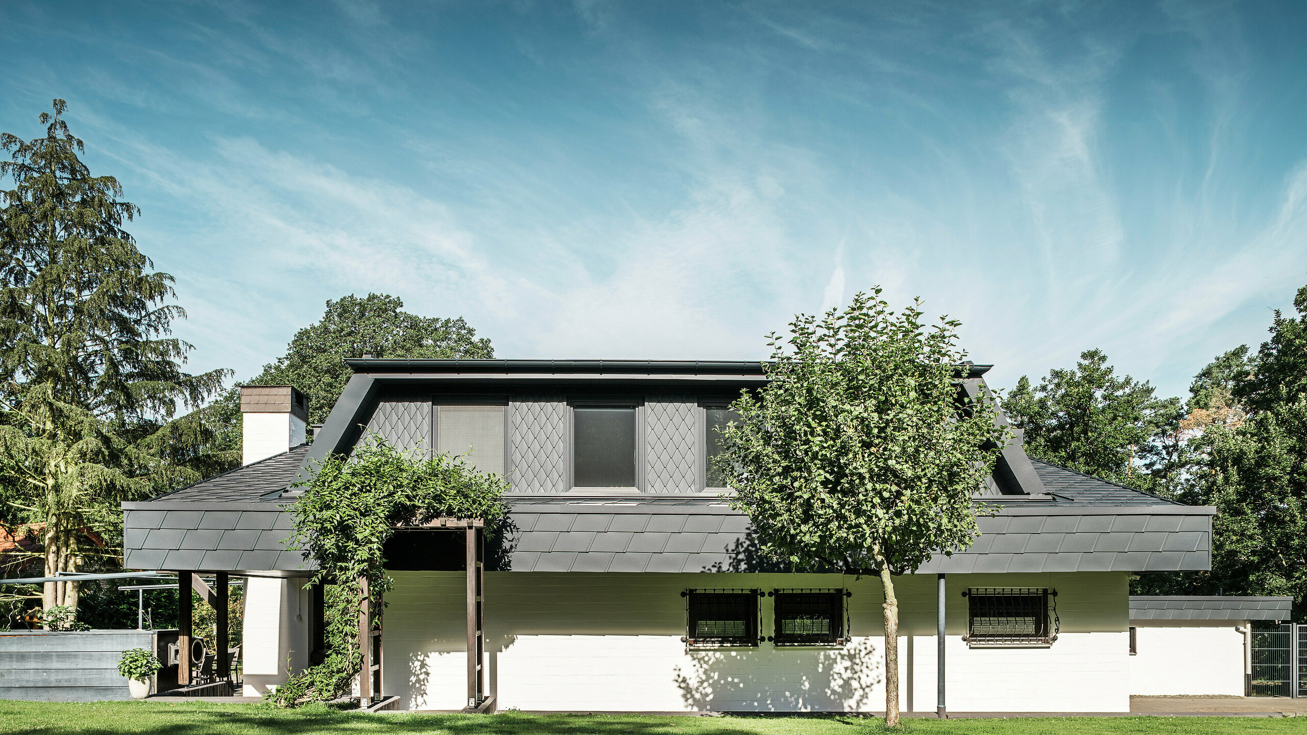 Fasadeudsnit mod haven på et enfamiliehus i Mechernich med en stor trapezformet kvist, dækket med små PREFA-tagromberer P.10 antracit. Taget er dækket med PREFA-tagspån DS.19 i samme farve. Facades på underetagen er malet hvid, og en beplantet pergola fuldender helhedsindtrykket i haveområdet. Træer og buske indrammer udsigten og sikrer en harmonisk integration i naturen.