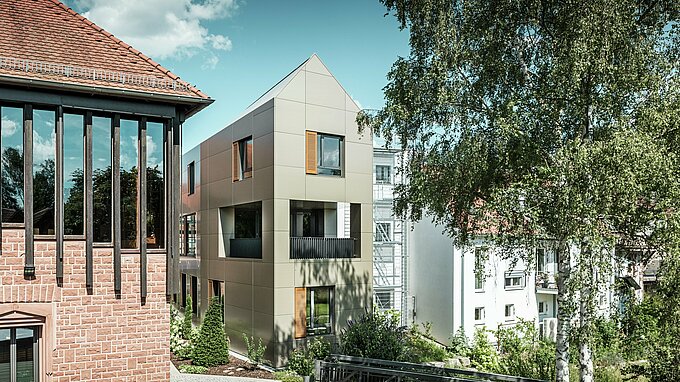 Das Bild zeigt das moderne Rathaus in Alzenau, dessen Fassade aus Aluminium-Verbundplatten von PREFA in Bronze besteht. Der spitze Dachbereich und die klar strukturierten Fenster mit Holzjalousien verleihen dem Gebäude eine elegante und zeitgenössische Ästhetik. Im Vordergrund ist ein grüner Bereich mit einem gepflasterten Weg und Bäumen zu sehen, der das urbane Design des Gebäudes harmonisch ergänzt.