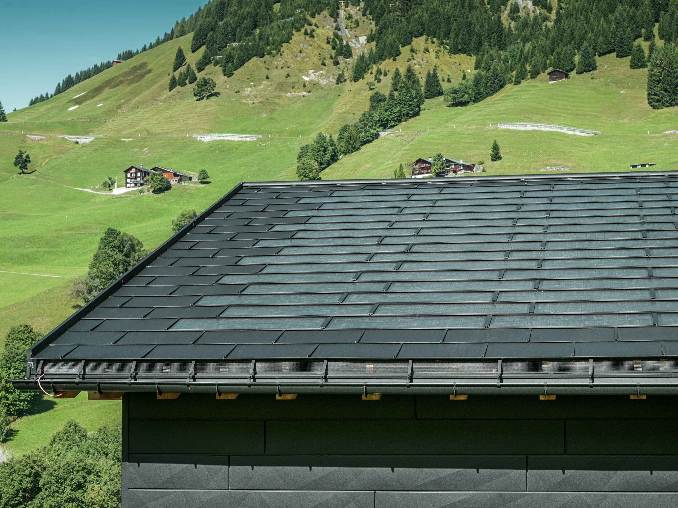 Nærbillede af tagskægget på et moderne enfamiliehus i de alpine omgivelser i Ascharina i Schweiz. Taget er dækket med den innovative PREFA-solcelletagplade og den traditionelle tagplade R.16, begge i P.10 sort. PREFA halvrund tagrende og PREFA nedløbsrør sørger for pålidelig tagafvanding. Nederst på billedet ses de karakteristiske PREFA Siding.X facadepaneler. De klare linjer i aluminiumsprodukterne understreger det moderne design midt i et malerisk bjerglandskab.