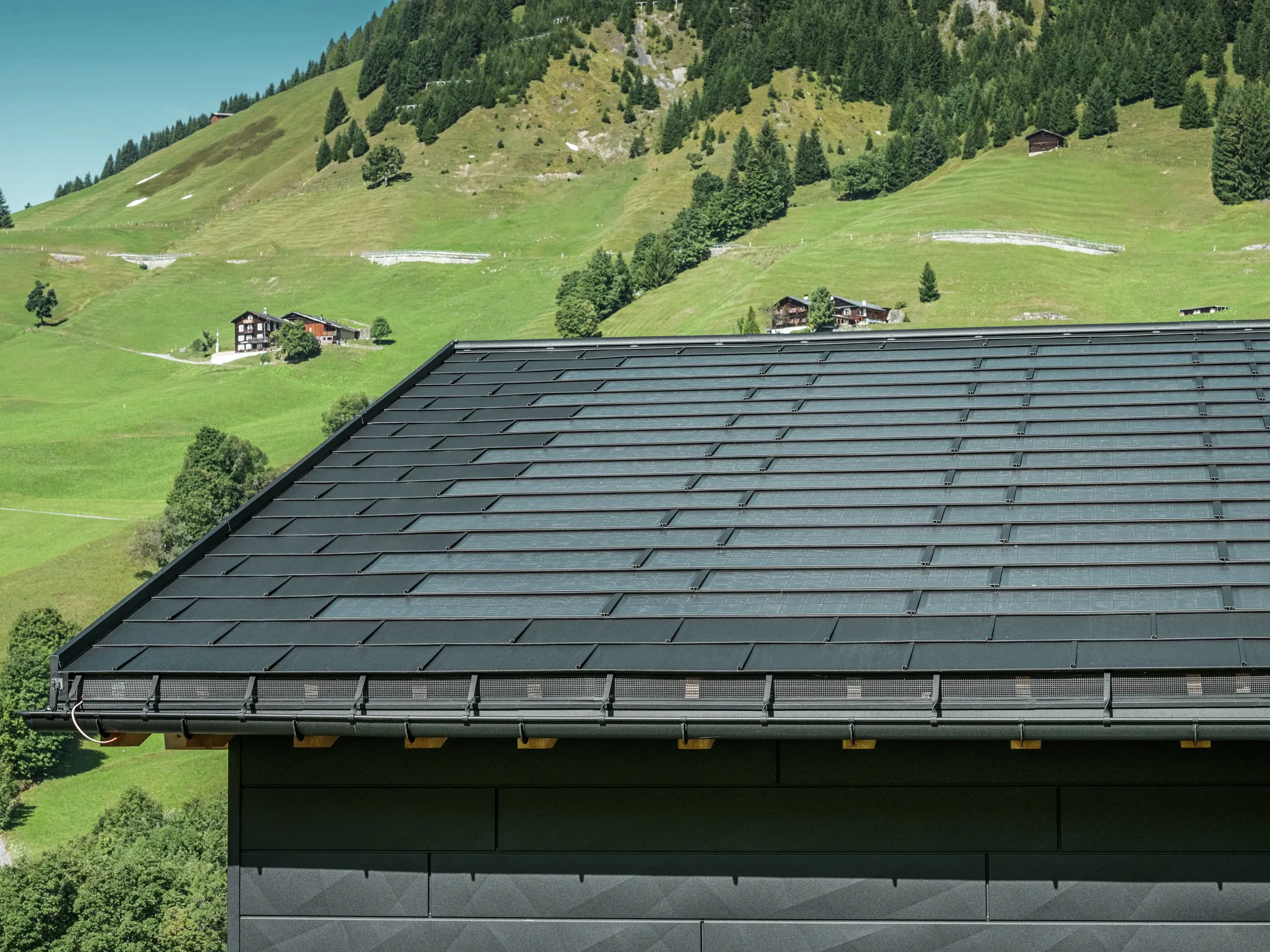 Nærbillede af tagskægget på et moderne enfamiliehus i de alpine omgivelser i Ascharina i Schweiz. Taget er dækket med den innovative PREFA-solcelletagplade og den traditionelle tagplade R.16, begge i P.10 sort. PREFA halvrund tagrende og PREFA nedløbsrør sørger for pålidelig tagafvanding. Nederst på billedet ses de karakteristiske PREFA Siding.X facadepaneler. De klare linjer i aluminiumsprodukterne understreger det moderne design midt i et malerisk bjerglandskab.