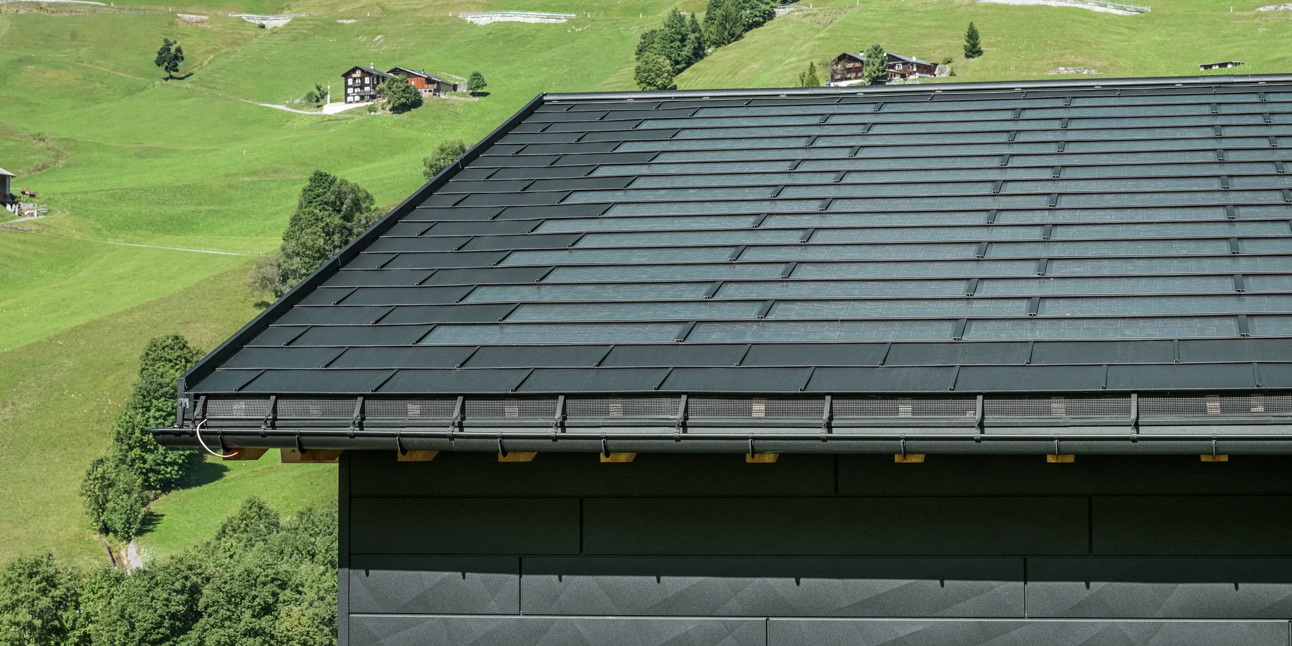 Nærbillede af tagskægget på et moderne enfamiliehus i de alpine omgivelser i Ascharina i Schweiz. Taget er dækket med den innovative PREFA-solcelletagplade og den traditionelle tagplade R.16, begge i P.10 sort. PREFA halvrund tagrende og PREFA nedløbsrør sørger for pålidelig tagafvanding. Nederst på billedet ses de karakteristiske PREFA Siding.X facadepaneler. De klare linjer i aluminiumsprodukterne understreger det moderne design midt i et malerisk bjerglandskab.