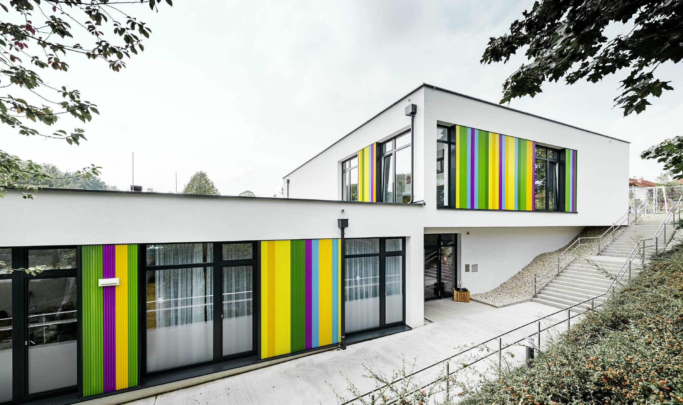 Farverige facadeelementer fra PREFA til børnehaven i Hargelsberg. Bygningen har et fladt tag og store, gulv til loft-vinduer.