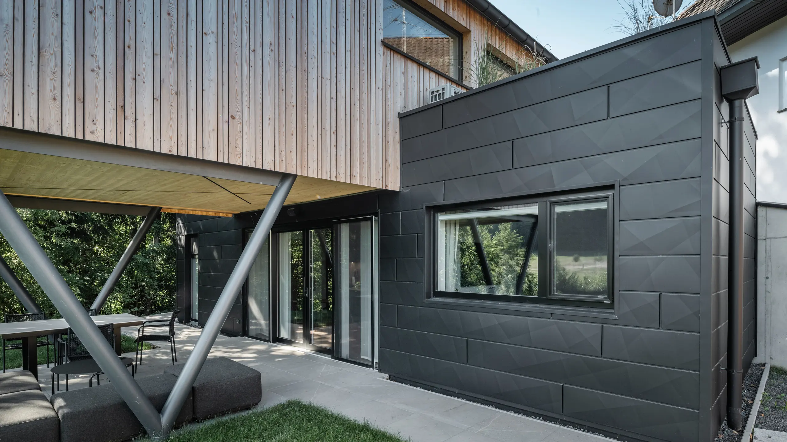 Arkitektonisk højdepunkt: ›Woodstock Chalet 5‹ i St. Martin am Innkreis, Oberösterreich. Den moderne bryllupsbolig fremhæves ved brug af antracitfarvede PREFA Siding.X-facadepaneler. Aluminiumselementerne danner en stærk kontrast til de lyse træpaneler og de metalliske bærende konstruktioner. De mørke vinduesrammer passer harmonisk ind i facadens design, hvilket giver bygningen et elegant udseende. Dette billede viser den æstetiske balance mellem naturlig træstruktur og den industrielle charme ved aluminiumsfacaden, som er kendetegnende for designet af moderne bygninger.