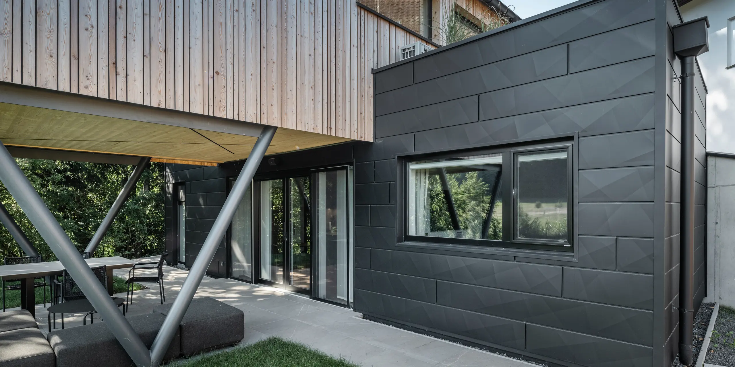 Arkitektonisk højdepunkt: ›Woodstock Chalet 5‹ i St. Martin am Innkreis, Oberösterreich. Den moderne bryllupsbolig fremhæves ved brug af antracitfarvede PREFA Siding.X-facadepaneler. Aluminiumselementerne danner en stærk kontrast til de lyse træpaneler og de metalliske bærende konstruktioner. De mørke vinduesrammer passer harmonisk ind i facadens design, hvilket giver bygningen et elegant udseende. Dette billede viser den æstetiske balance mellem naturlig træstruktur og den industrielle charme ved aluminiumsfacaden, som er kendetegnende for designet af moderne bygninger.