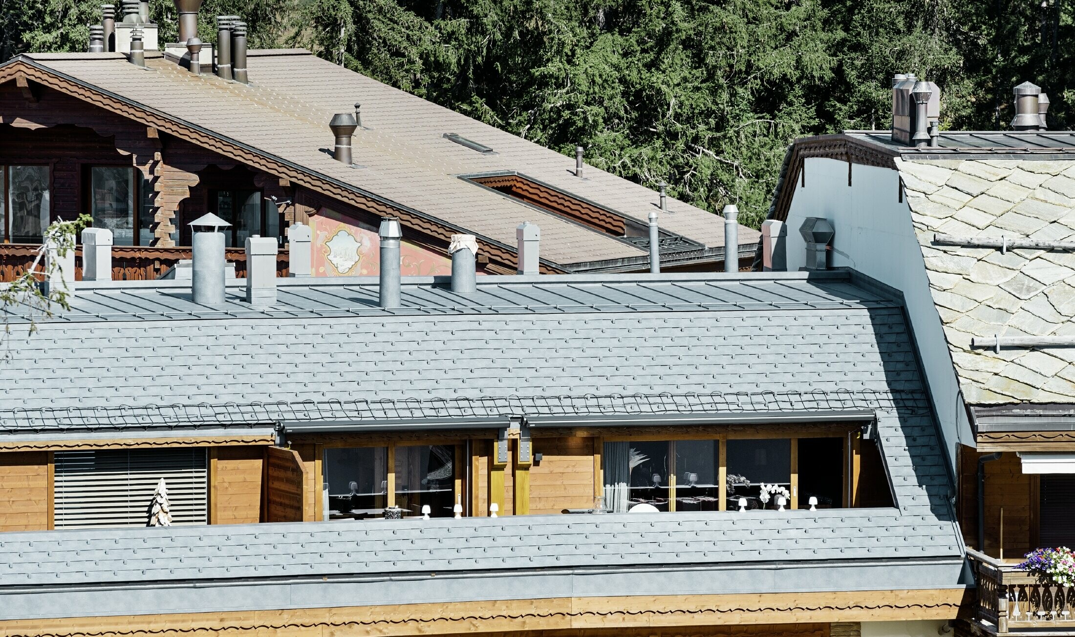 Flerfamiliehus i Crans-Montana med bjerge i baggrunden og en facade med fantasifulde træelementer med en PREFA aluminium tagspån-tag i stengrå