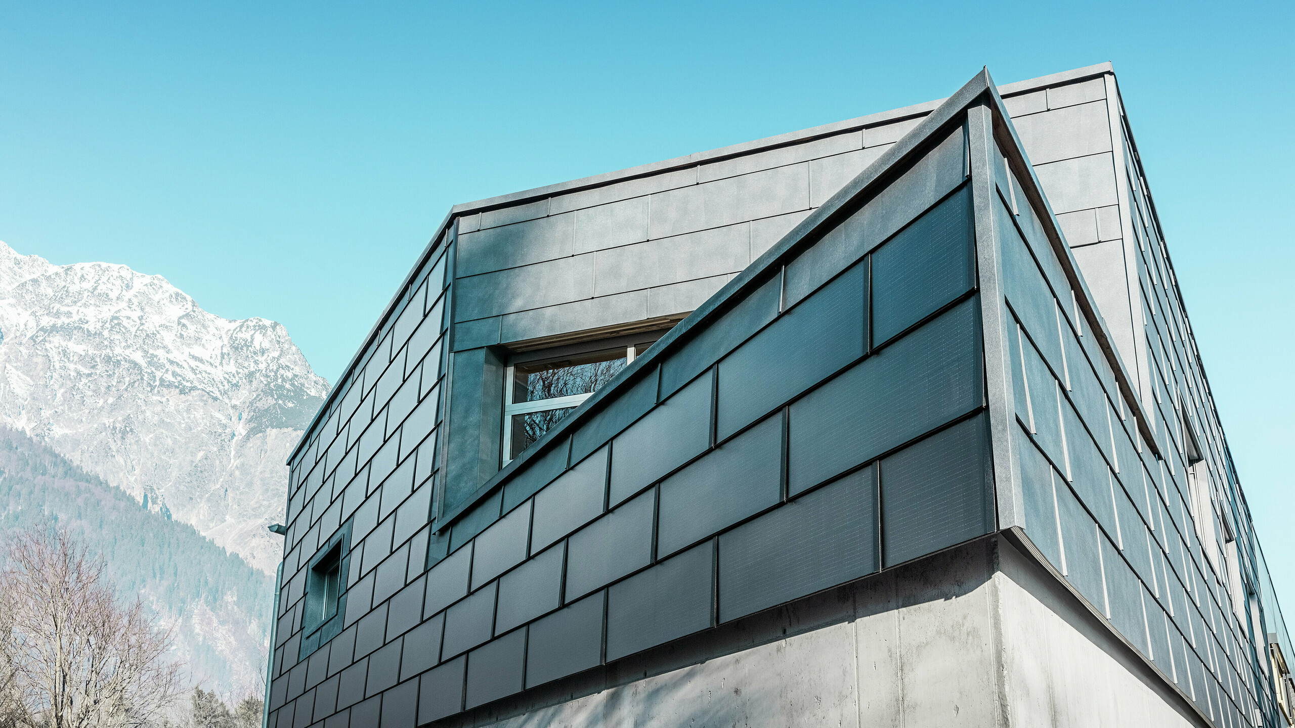 Fassade des Firmengebäudes der Spenglerei RÜMA in Vorarlberg mit eleganten, grauen PREFA Solardachplatten und PREFA Fassadenpaneelen FX.12, kombiniert mit Sichtbeton, vor klar blauem Himmel und Bergkulisse. Die spitze Gebäudeecke unterstreicht die auffallende Architektur.