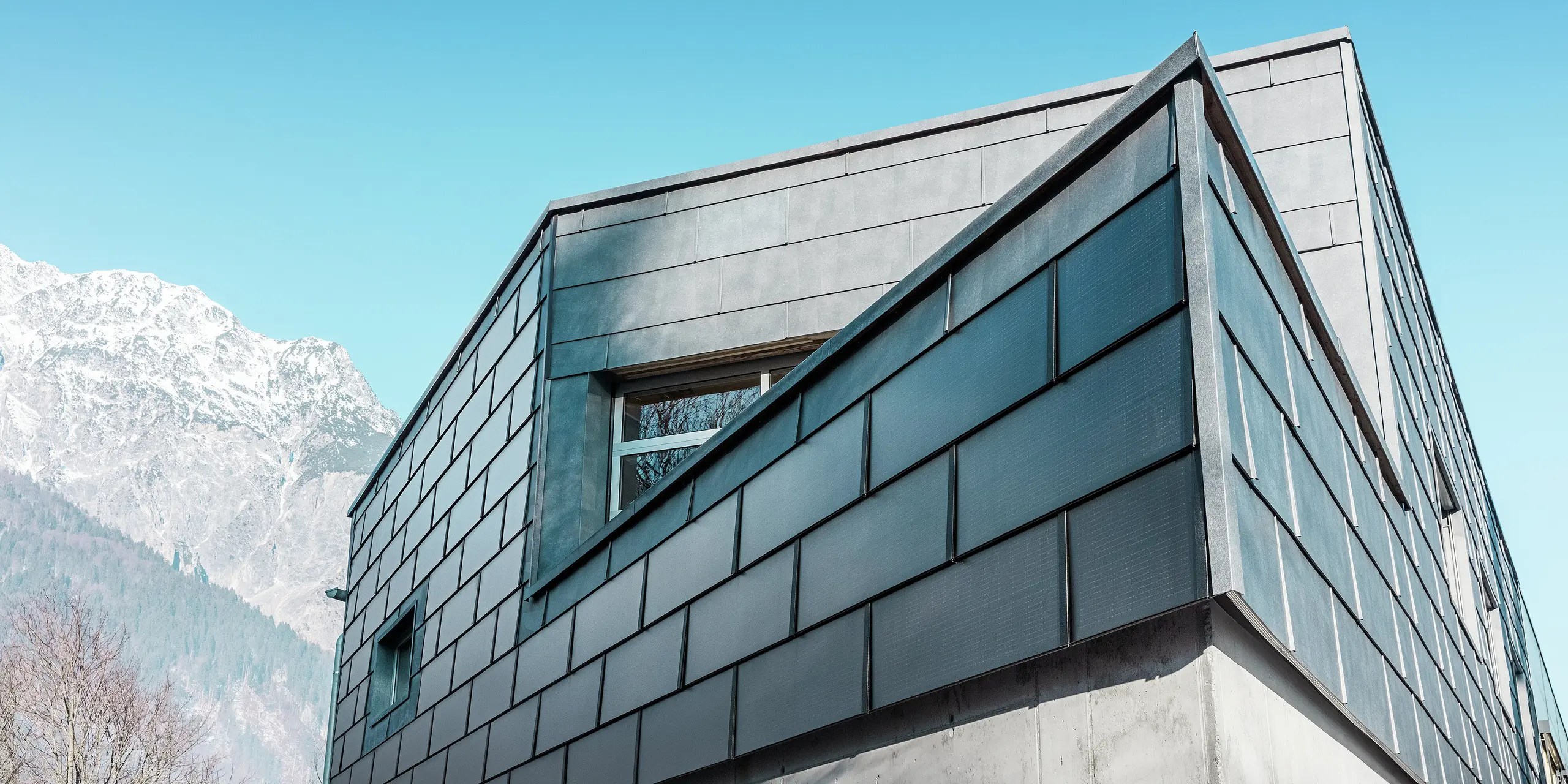 Facade på firmaet RÜMA's bygning i Vorarlberg med elegante, grå PREFA-solcelletagplader og PREFA facadepanel FX.12, kombineret med synlig beton, foran en klar blå himmel og bjergkulisse. Den spidse hjørne af bygningen understreger den iøjnefaldende arkitektur.
