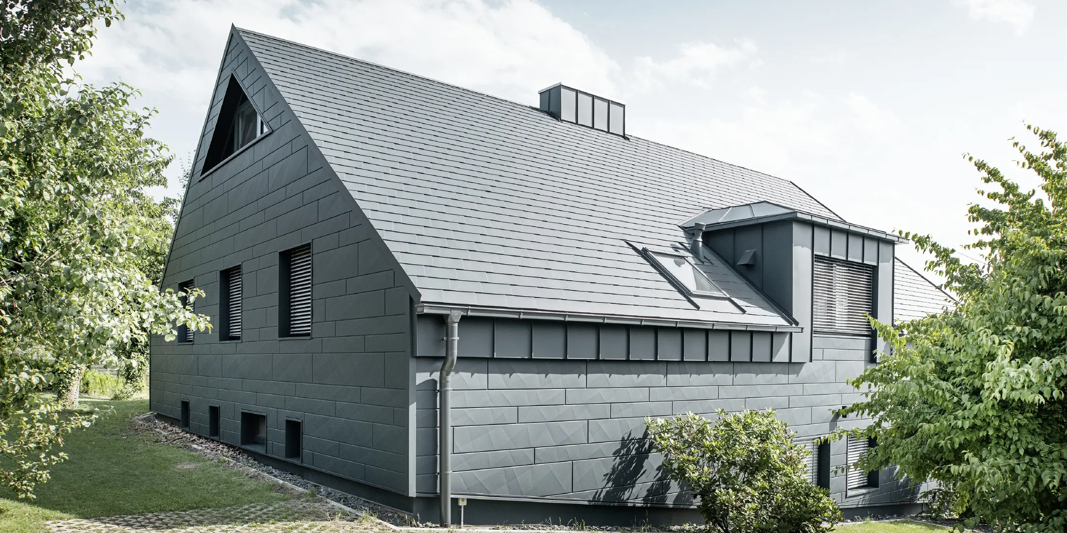 Stort enfamiliehus med sadeltag og mange vinduer. Hele huset er beklædt med en aluminiumsbeklædning i antracit, på taget er der anvendt PREFA aluminiums tagspån, og på facaden er der anvendt Siding.X i antracit.