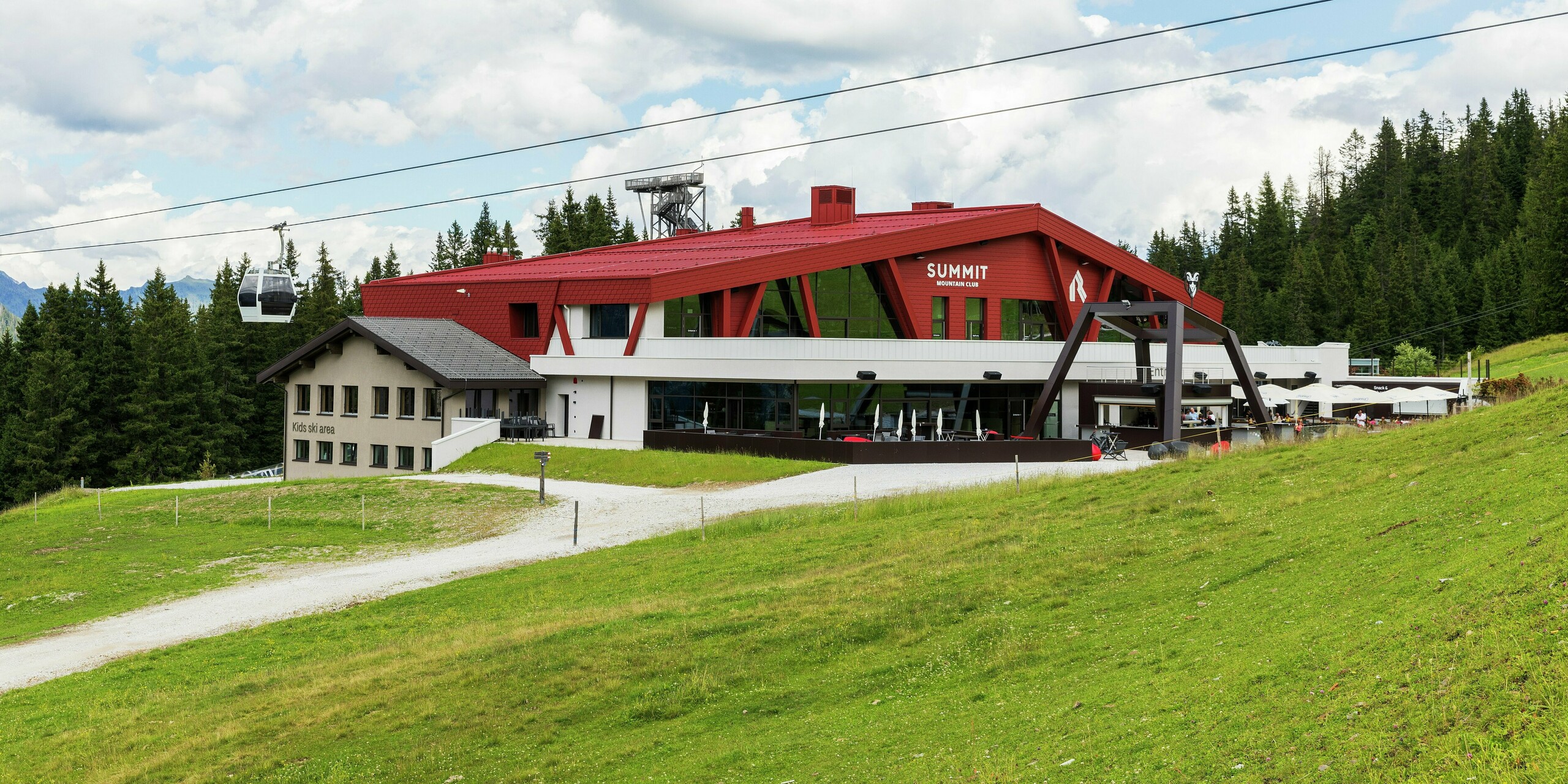 Der Summit Mountain Club in Ratschings präsentiert sich mit einem markanten Dach und einer Fassade in der Farbe P.10 Oxydrot. Die moderne Architektur fügt sich perfekt in die alpine Landschaft ein. Die auffällige Struktur in Kombination mit großen Fensterflächen zieht sofort die Blicke auf sich. Im Vordergrund ist die Zugangsstraße sichtbar. Das mit PREFA Wandschindeln und PREFALZ-Stehfalzbahnen verkleidete Gebäude bietet nicht nur ästhetische, sondern auch funktionale Lösungen, die das raue Bergklima problemlos meistern.