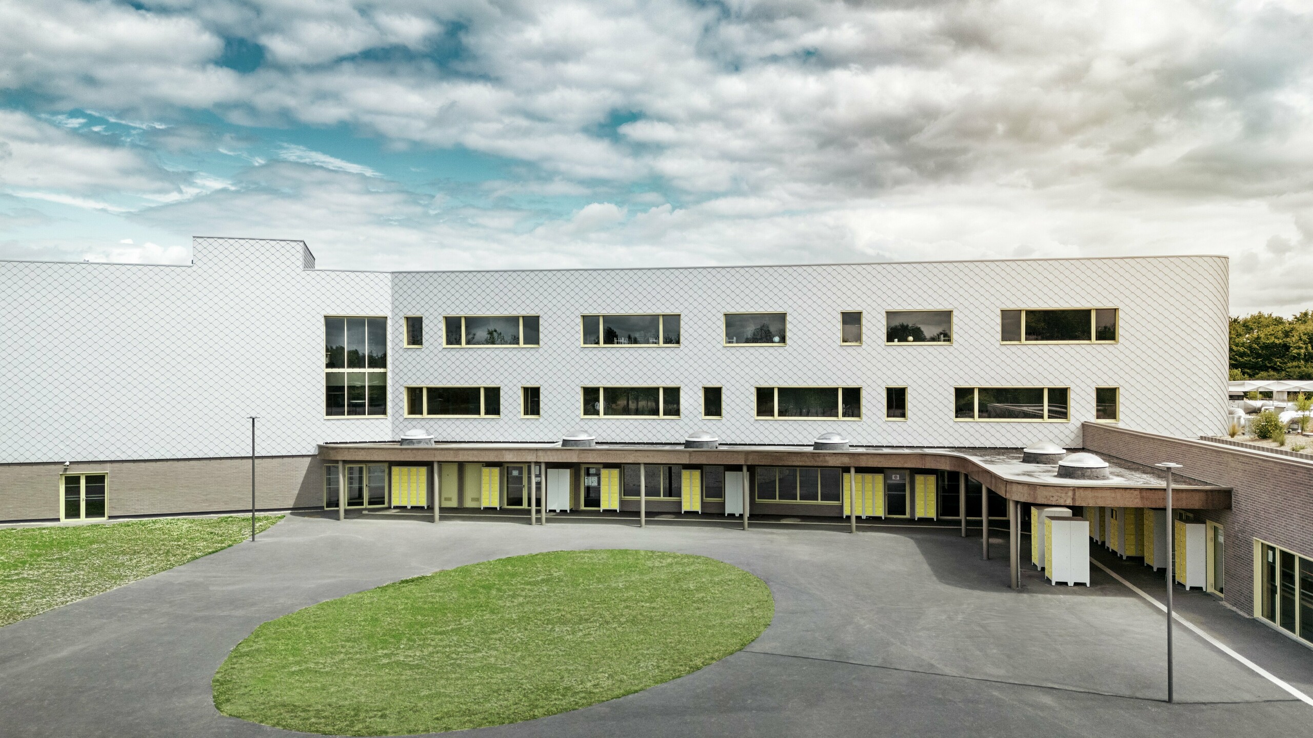 Rückseite des Collège Léontine Dolivet mit großem Schulhof und klarer Architektur. Die Fassade besteht aus silbermetallic PREFA Wandrauten, die für eine moderne, elegante Optik sorgen. Mehrere Fensterreihen und gelbe Türen lockern die Struktur auf, während der asphaltierte Hof mit zentraler Grünfläche viel Platz für Pausen bietet. Die langlebige Aluminiumfassade kombiniert Leichtigkeit, Witterungsbeständigkeit und Design – ein funktionaler und ästhetischer Eyecatcher in Cesson-Sévigné nahe Rennes.