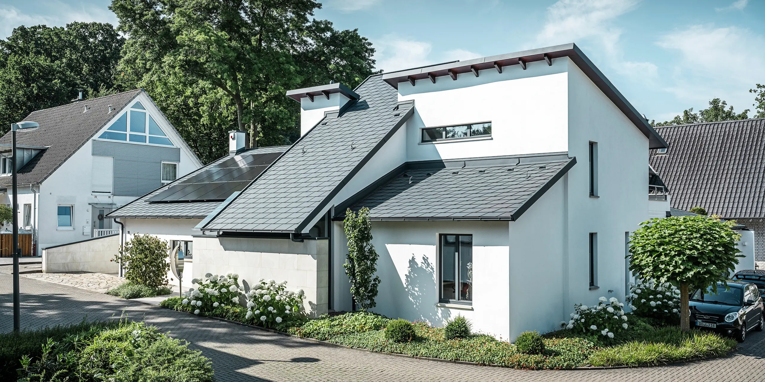 Moderne enfamiliehus med hvid pudset facade og mørkt PREFA-skråtag af aluminium. På taget ses integrerede solcellemoduler og en tagafvanding af aluminium i antracit. Bygningen ligger i et velholdt boligkvarter med beplantede forhaver, asfalteret indkørsel og omkringliggende enfamiliehuse.