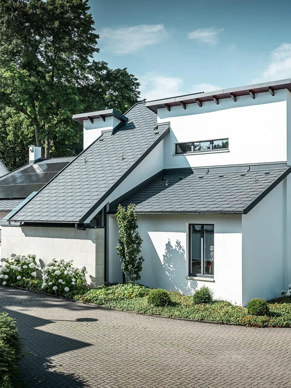 Moderne enfamiliehus med hvid pudset facade og mørkt PREFA-skråtag af aluminium. På taget ses integrerede solcellemoduler og en tagafvanding af aluminium i antracit. Bygningen ligger i et velholdt boligkvarter med beplantede forhaver, asfalteret indkørsel og omkringliggende enfamiliehuse.