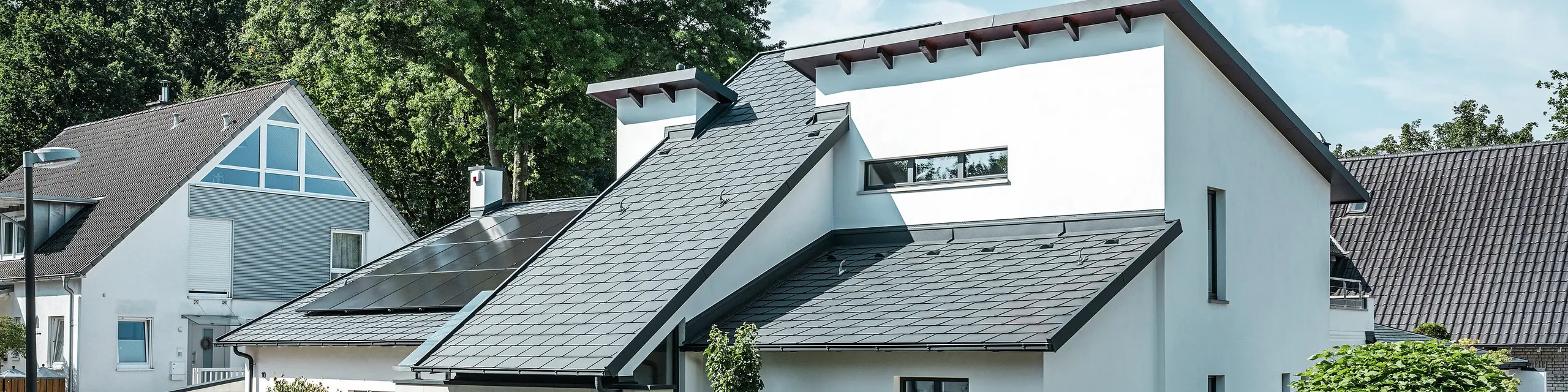 Moderne enfamiliehus med hvid pudset facade og mørkt PREFA-skråtag af aluminium. På taget ses integrerede solcellemoduler og en tagafvanding af aluminium i antracit. Bygningen ligger i et velholdt boligkvarter med beplantede forhaver, asfalteret indkørsel og omkringliggende enfamiliehuse.