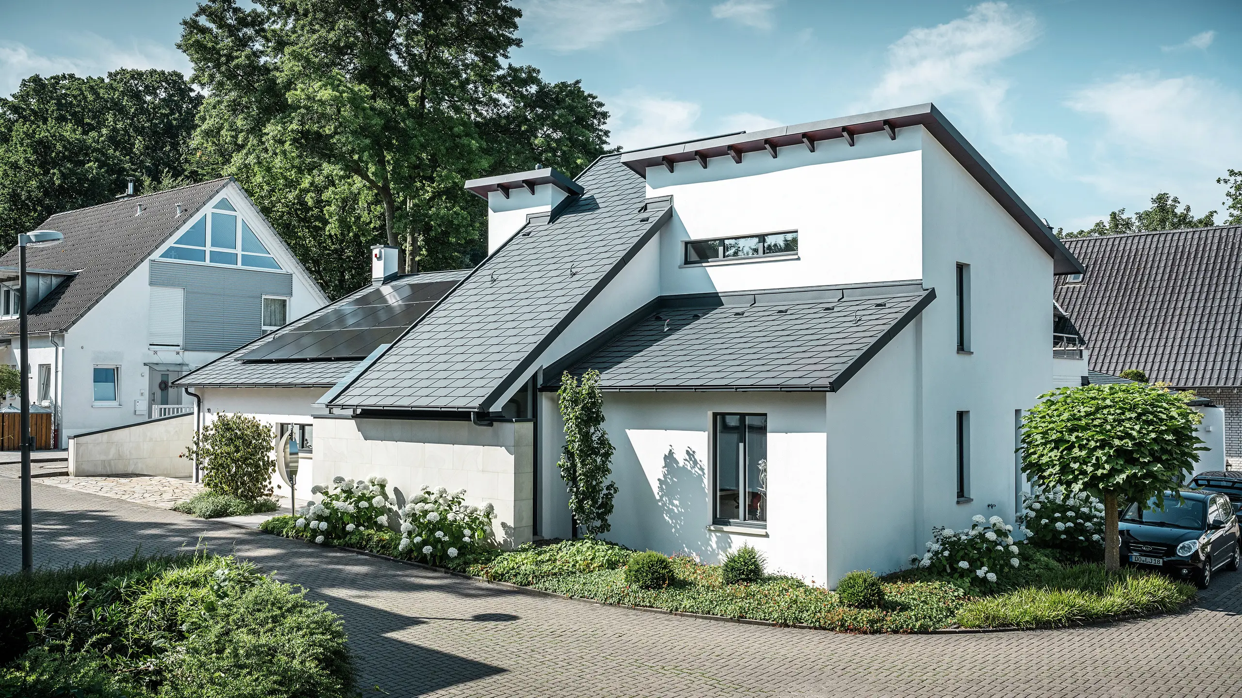 Moderne enfamiliehus med hvid pudset facade og mørkt PREFA-skråtag af aluminium. På taget ses integrerede solcellemoduler og en tagafvanding af aluminium i antracit. Bygningen ligger i et velholdt boligkvarter med beplantede forhaver, asfalteret indkørsel og omkringliggende enfamiliehuse.