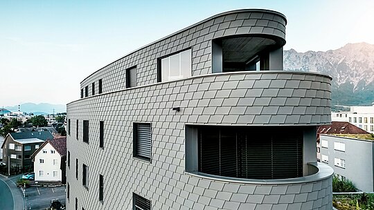 Detailreiche Schrägansicht des Bernerhauses in Buchs mit weich gerundeter Ecke und durchlaufenden Balkonbändern. Die Fassade aus PREFA Wandschindeln in P.10 Hellgrau legt sich wie eine präzise verarbeitete Aluminiumhaut über die dreieckige Kubatur und erzeugt ein lebendiges, schuppenartiges Muster. Die homogene Oberfläche betont die fließenden Radien und das Spiel von Licht und Schatten. Dank des leichten, witterungsbeständigen und langlebigen PREFA Aluminiums entsteht eine wartungsarme Gebäudehülle mit markanter Fernwirkung vor alpiner Kulisse.