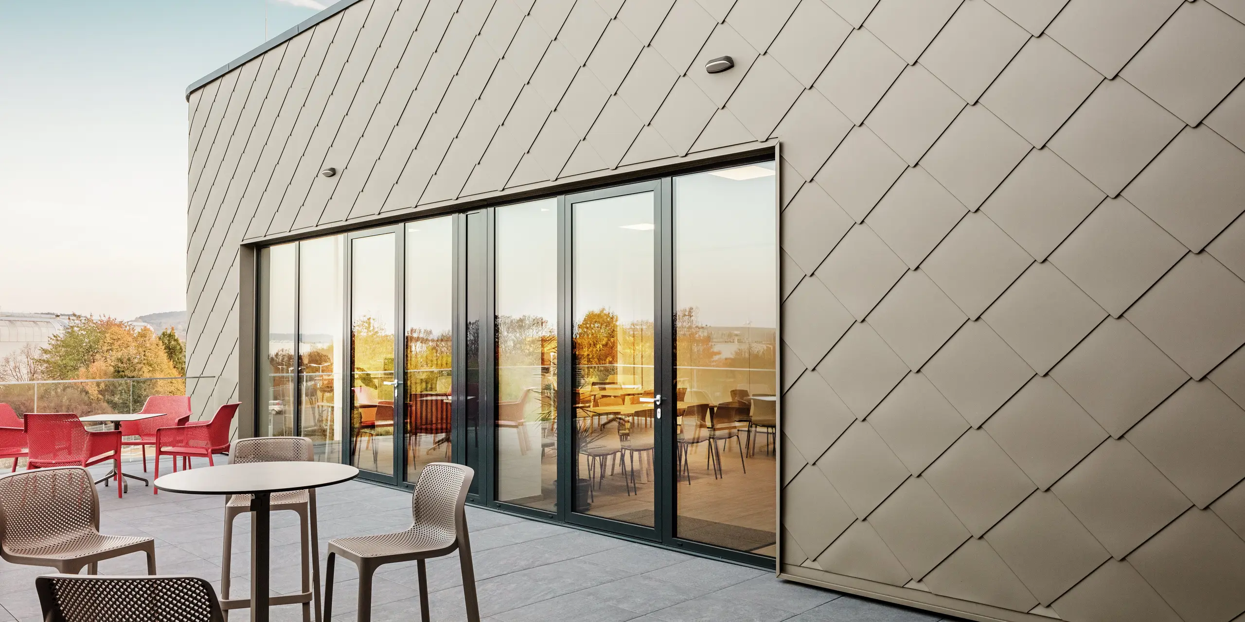 Facaden på det nye PREFA-anlæg i Törökbálint imponerer med de elegante PREFA-vægromber 44 × 44 i P.10 prefabronze. Den præcist monterede aluminiumsfacade sikrer et moderne, dynamisk udseende og tilbyder samtidig maksimal beskyttelse mod vejrliget. Den store glasfront forbinder det indvendige og udvendige rum og skaber en lys og indbydende atmosfære på den store terrasse.