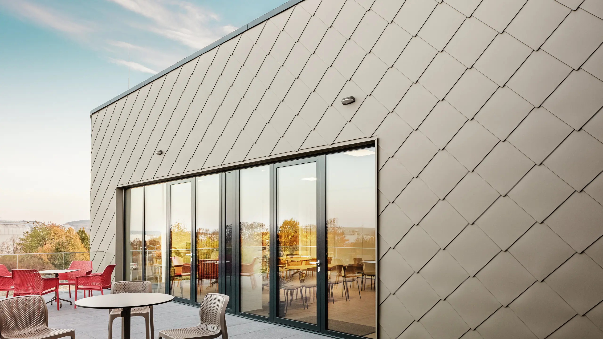 Facaden på det nye PREFA-anlæg i Törökbálint imponerer med de elegante PREFA-vægromber 44 × 44 i P.10 prefabronze. Den præcist monterede aluminiumsfacade sikrer et moderne, dynamisk udseende og tilbyder samtidig maksimal beskyttelse mod vejrliget. Den store glasfront forbinder det indvendige og udvendige rum og skaber en lys og indbydende atmosfære på den store terrasse.