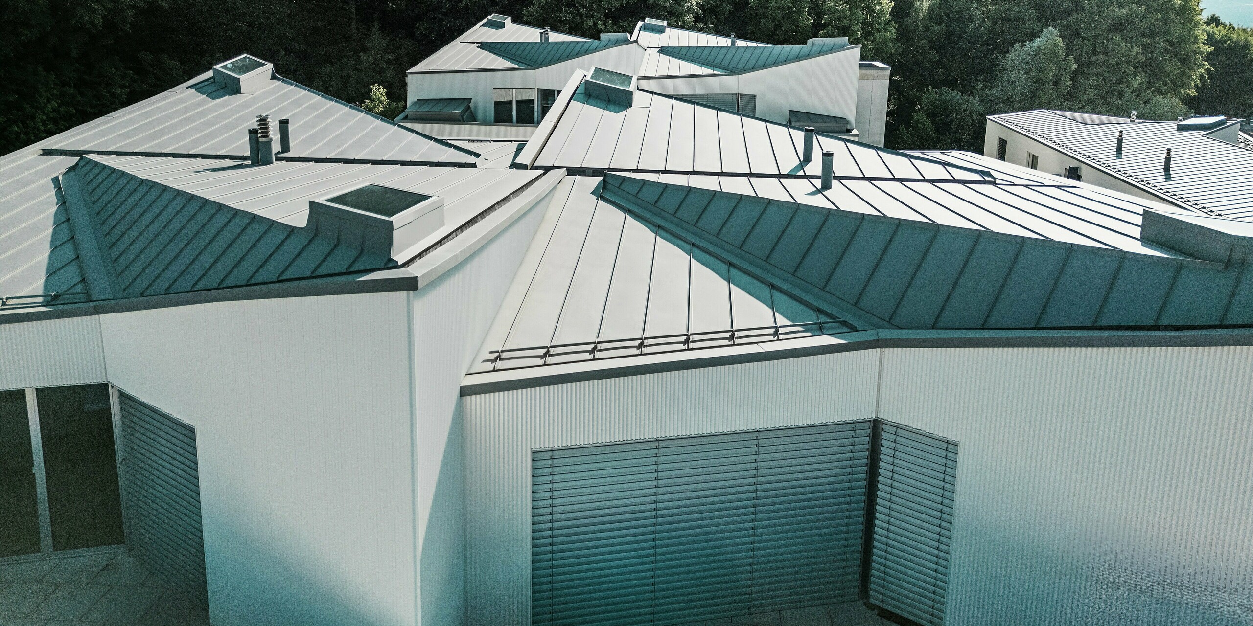 Panoramaaufnahme der außergewöhnlich gefalteten PREFALZ Dächer in Patinagrau über einer Gruppe moderner Tiny Homes. Die präzise verarbeiteten Stehfalzflächen erzeugen ein lebendiges Spiel aus Licht und Schatten. Das langlebige PREFA Aluminium schützt zuverlässig vor Wind und Wetter und ermöglicht architektonisch anspruchsvolle Dachformen auf engstem Raum.