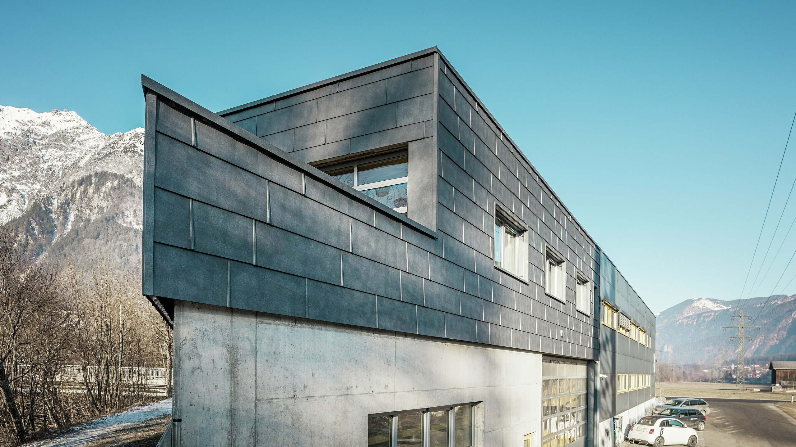 Die Vorderseite des Firmengebäudes der Spenglerei RÜMA in Vorarlberg. Es verfügt über eine markante Fassade aus grauen PREFA FX.12-Fassadenpaneelen aus Aluminium und einen sichtbaren Betonunterbau. Es ist in eine alpine Landschaft mit Bergen und blauem Himmel eingebettet. Auf dem Parkplatz vor dem Gebäude ist ein ladendes Elektroauto zu sehen, das die Nachhaltigkeit des Unternehmens unterstreicht.