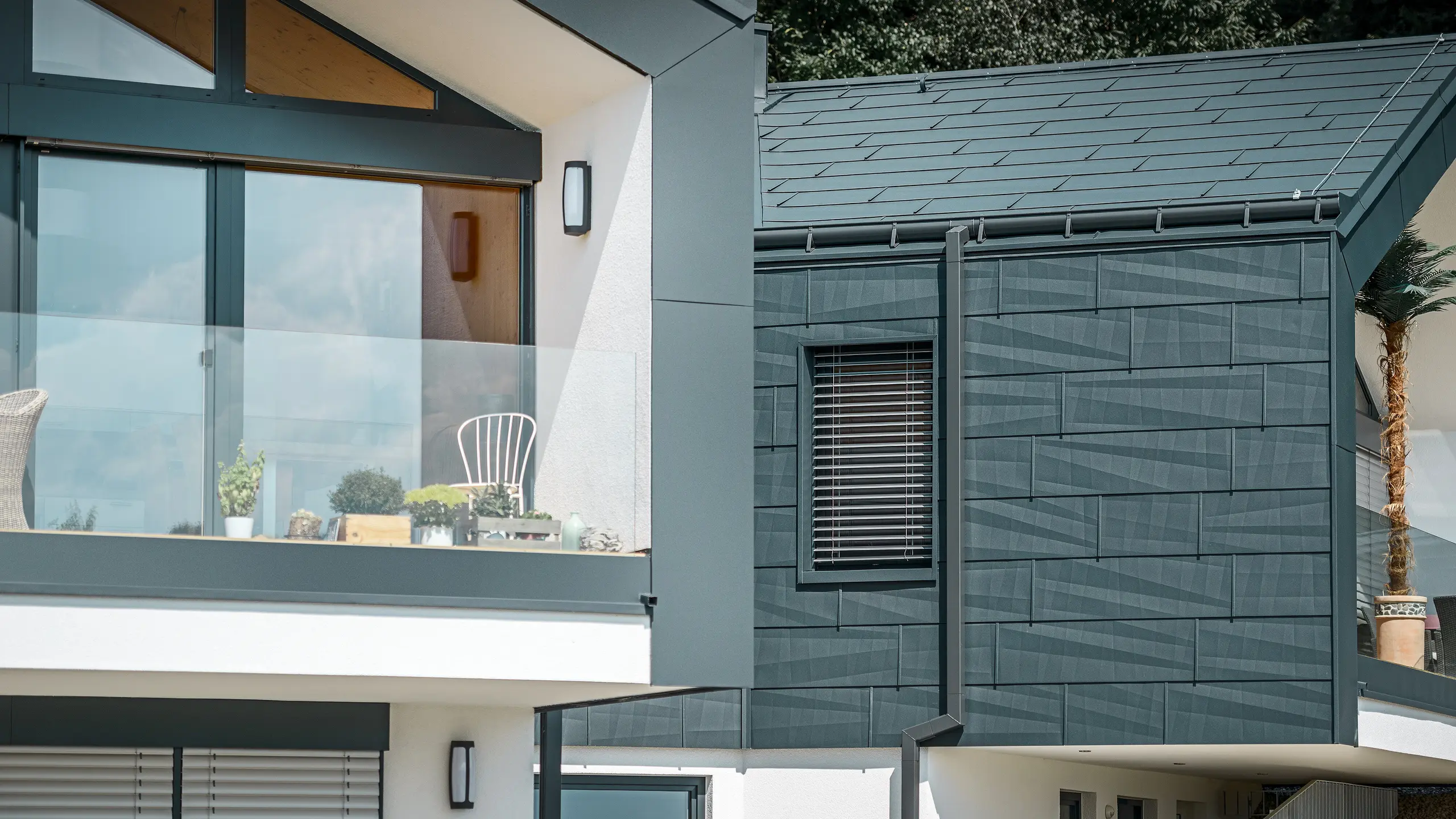 Billedet viser et moderne dobbelthus med en stilfuld facade i aluminium. Facaden er holdt i en mørk antracitfarve og har en struktureret overflade takket være PREFA tag- og facadepanel FX.12. På taget er der monteret en fodrende og et kvadratrør som nedløbsrør fra PREFA. På øverste etage ses en altan med glasgelænder, mens der på nederste etage er vinduer med lamelljalousier. Arkitekturen fremstår klar og minimalistisk, og i baggrunden ses træer, der skaber en naturlig atmosfære.