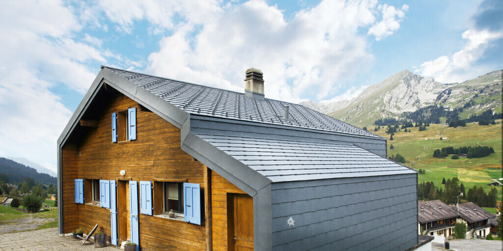 Hus i bjergene i Schweiz med træfacade kombineret med PREFA-tagrombe og -vægrombe i stengrå