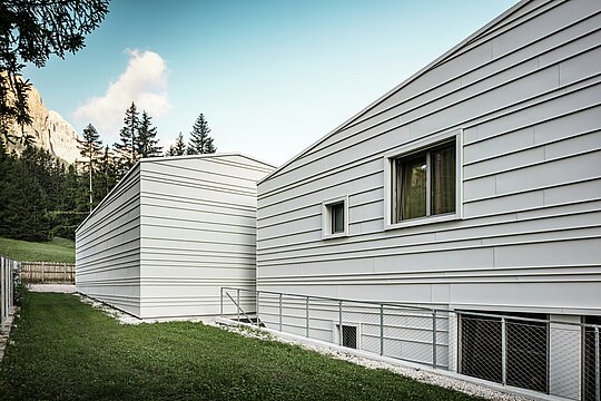 Stražnja strana vojarne „Tempesti“ u Corvari pruža dojmljivu perspektivu: vide se šumska stabla, blistavo plavo nebo i suncem obasjani planinski vrhovi. Svijetla aluminijska fasada s jasnom strukturom odražava tehničku sofisticiranost PREFALZ-a u boji P.10 Prefaweiß. Horizontalne aluminijske trake reflektiraju svjetlost, pružaju optimalnu zaštitu od vremenskih utjecaja i skladno se uklapaju u alpski krajolik. Pravi primjer dugotrajne i moderne vojne arhitekture!