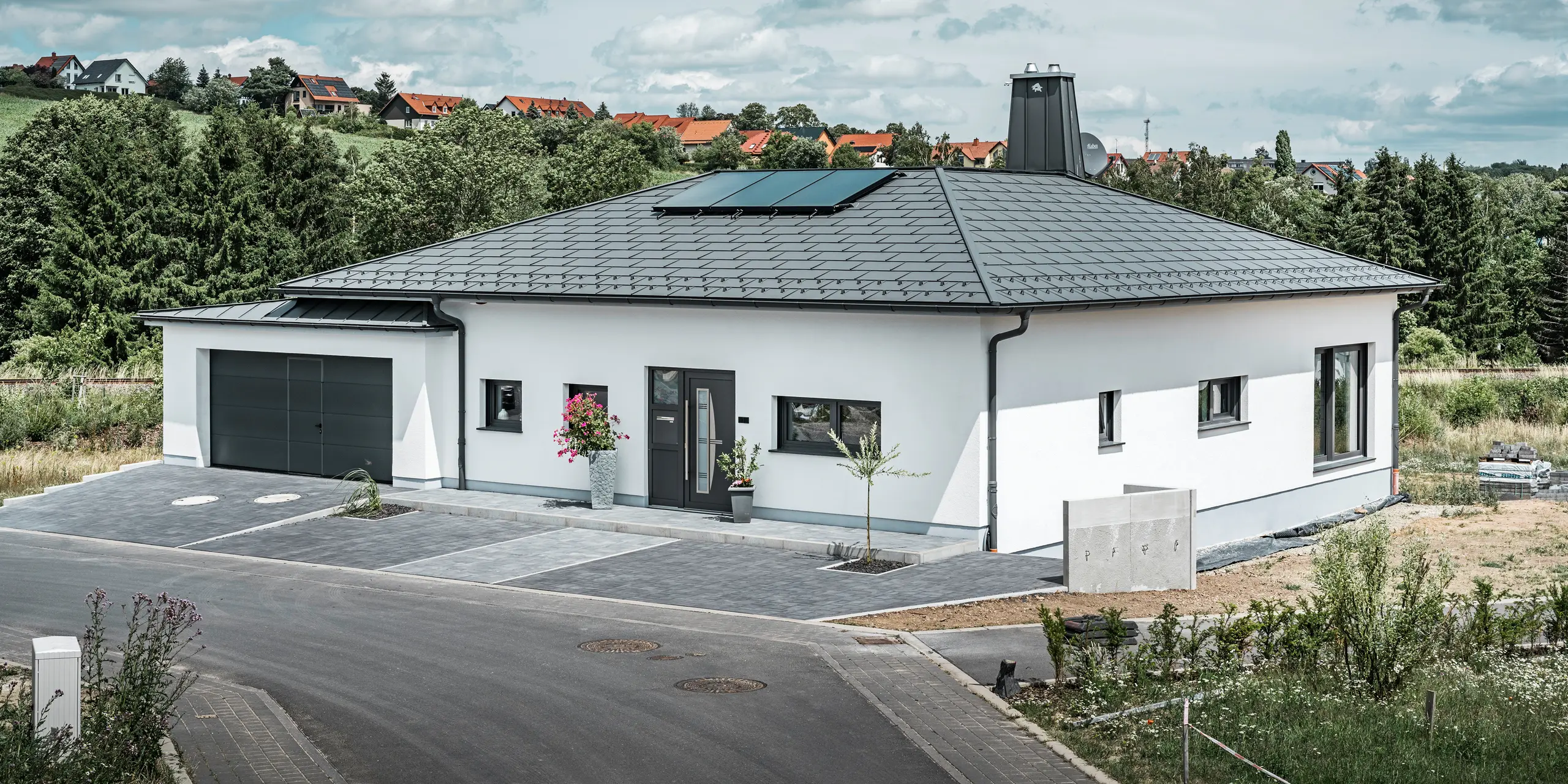Elegant enfamiliehus i Ilmenau, Thüringen, med et højkvalitets PREFA-tag i farven P.10 mørk grå, udstyret med den robuste tagplade R.16. Garagen harmonerer i farve og stil takket være brugen af PREFALZ. Ejendommens blikfang er det funktionelt designede atrium og skorstenen, der er dekoreret med PREFA-tyren, som understreger PREFAs moderne design og innovative tagteknologi. Solcellepanelerne passer perfekt ind i husets bæredygtige koncept. Det gennemtænkte helhedskoncept for bungalowen afrundes af PREFA-tagafvandingssystemet, som også er holdt i mørk grå.