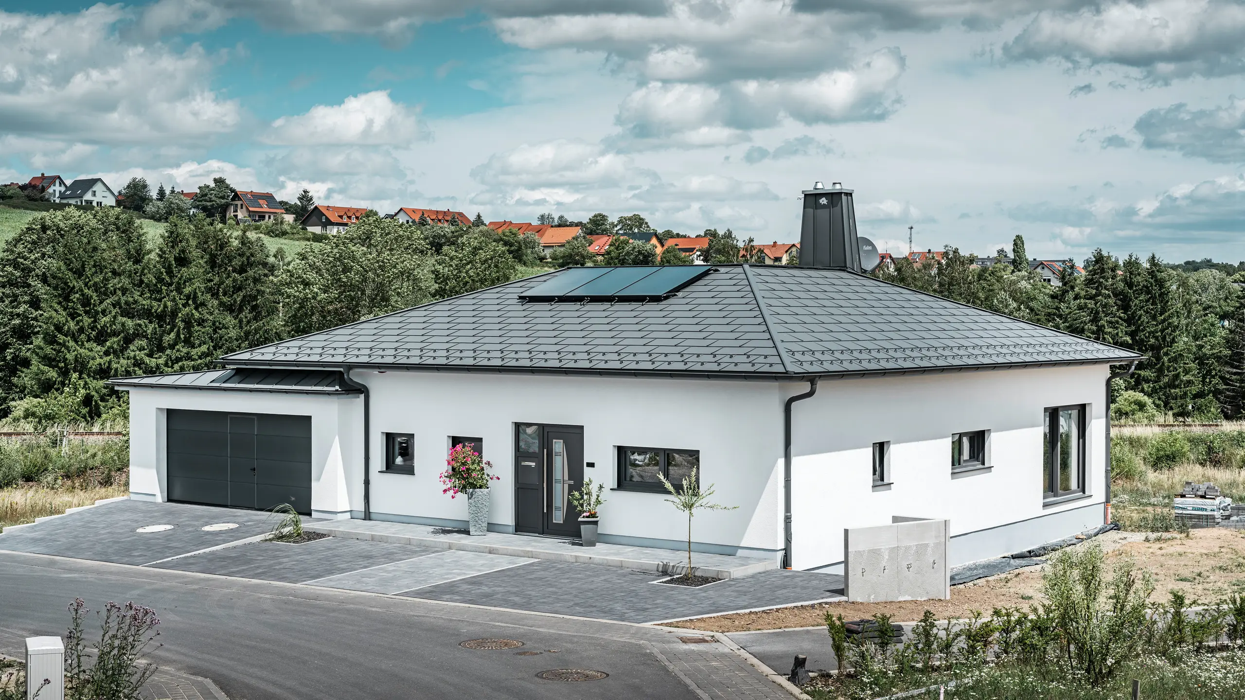 Elegant enfamiliehus i Ilmenau, Thüringen, med et højkvalitets PREFA-tag i farven P.10 mørk grå, udstyret med den robuste tagplade R.16. Garagen harmonerer i farve og stil takket være brugen af PREFALZ. Ejendommens blikfang er det funktionelt designede atrium og skorstenen, der er dekoreret med PREFA-tyren, som understreger PREFAs moderne design og innovative tagteknologi. Solcellepanelerne passer perfekt ind i husets bæredygtige koncept. Det gennemtænkte helhedskoncept for bungalowen afrundes af PREFA-tagafvandingssystemet, som også er holdt i mørk grå.