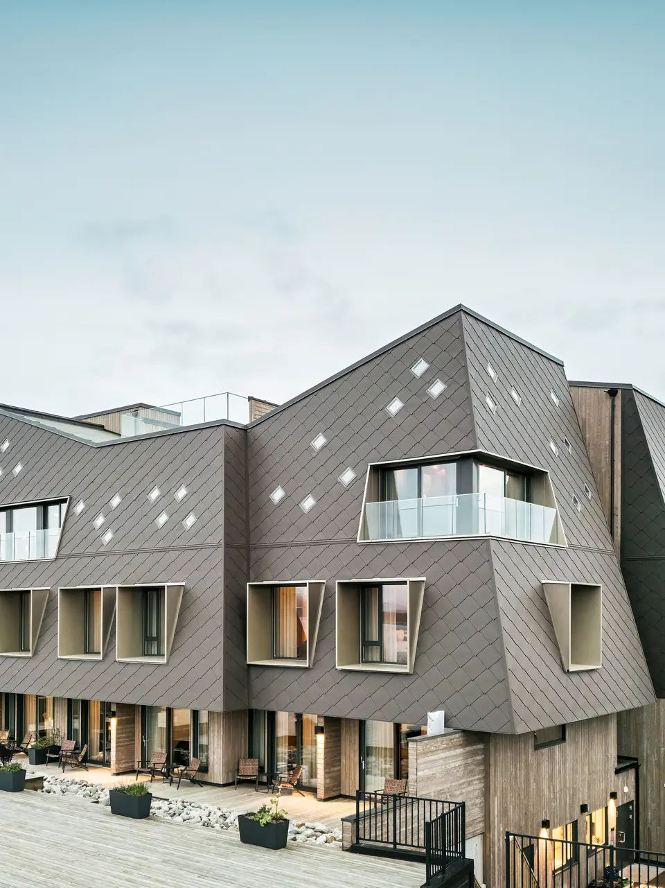 Blick auf die Terrasse des Hotels Beckerwyc House in Bekkjarvik, Norwegen. Das moderne Gebäude wird von einer einer markanten Aluminiumfassade aus PREFA Wandrauten 44 × 44 in Nussbraun geschmückt. Die futuristische Architektur mit schrägen Dachflächen und hervortretenden Fensterelementen wird durch die großzügigen Außenbereiche ergänzt. Die natürliche Farbgebung der Fassade passt sich harmonisch in die umgebende Landschaft ein und unterstreicht das zeitgemäße Design des Gebäudes.