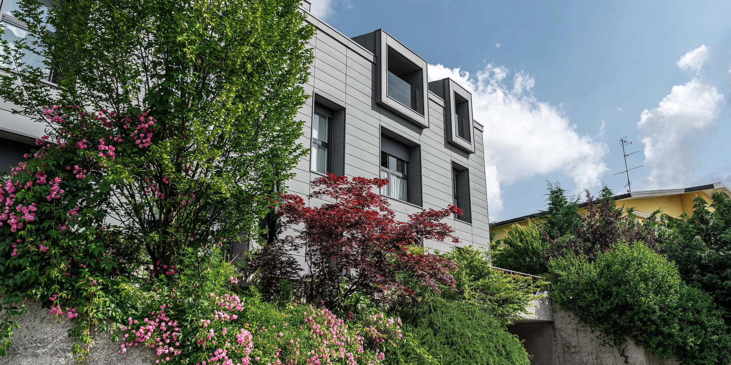 Moderne boligkompleks i Faloppio, Italien, med stilfuld facade af PREFA Sidings. De udkragede vinduer er et unikt arkitektonisk blikfang, mens de omgivende planter og blomster understreger bygningens naturlige skønhed. Den holdbare aluminiumsbeklædning fra PREFA er en æstetisk og funktionel løsning i al slags vejr.