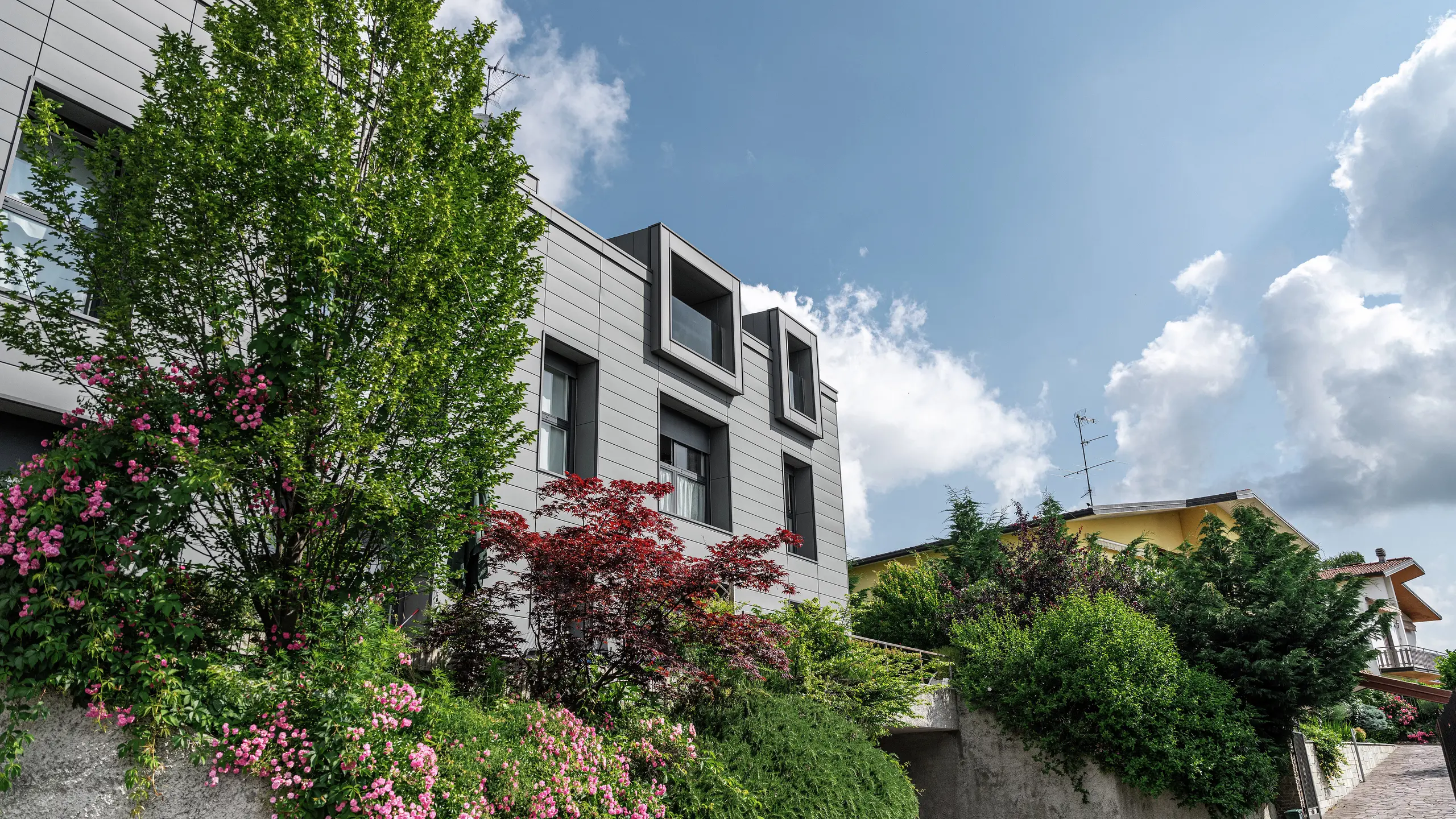 Moderne boligkompleks i Faloppio, Italien, med stilfuld facade af PREFA Sidings. De udkragede vinduer er et unikt arkitektonisk blikfang, mens de omgivende planter og blomster understreger bygningens naturlige skønhed. Den holdbare aluminiumsbeklædning fra PREFA er en æstetisk og funktionel løsning i al slags vejr.