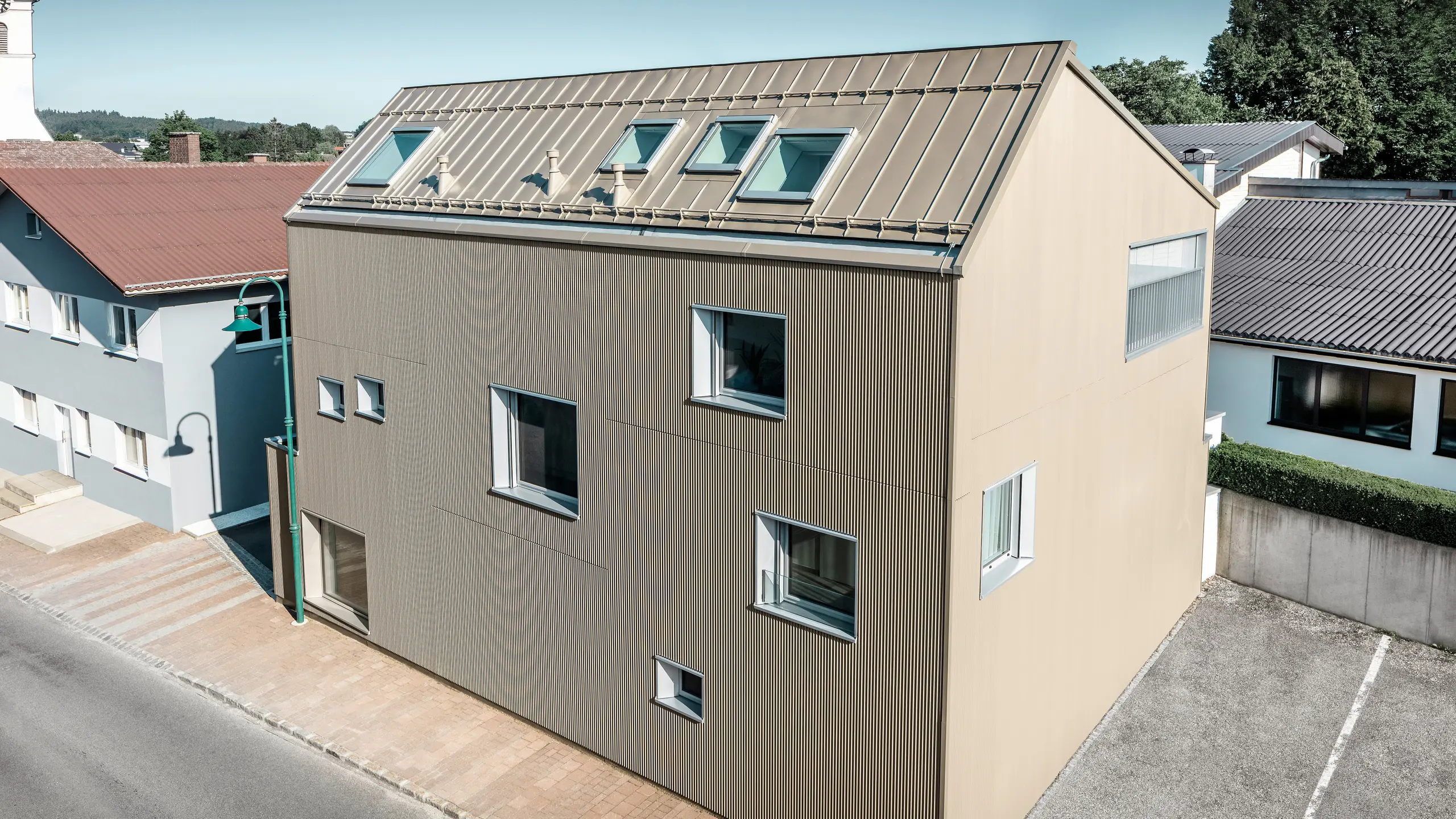 Fugleperspektiv på et enfamiliehus i Frankenburg, Østrig, der skiller sig ud med sin PREFA-facade i form af et takket profil og et PREFALZ-tag, begge i en afstemt bronzefarve. Facaden har en markant struktur, der fanger det naturlige lys og skaber et levende skuespil af skygger. Flere tagvinduer lader naturligt lys strømme ind og understreger den funktionelle og æstetiske integration af moderne arkitektoniske elementer. Huset passer med sit moderne design harmonisk ind i det typiske østrigske landsbymiljø og skaber samtidig en kontrast til de omkringliggende traditionelle bygninger.