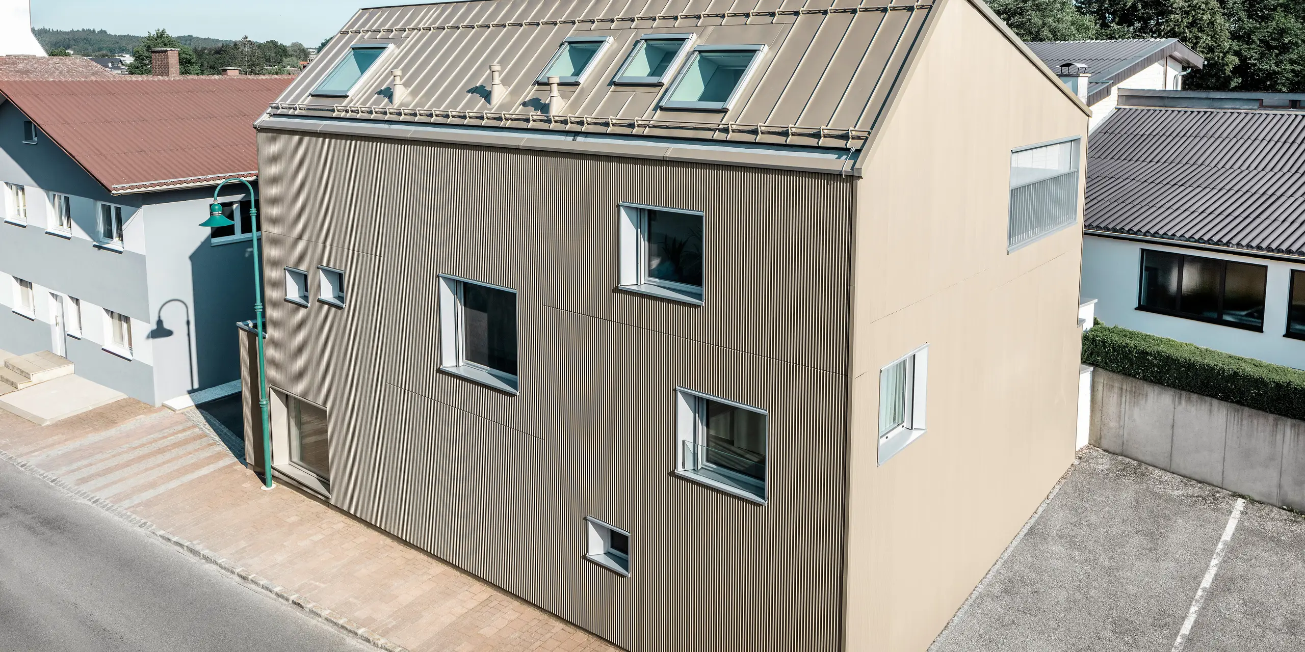 Fugleperspektiv på et enfamiliehus i Frankenburg, Østrig, der skiller sig ud med sin PREFA-facade i form af et takket profil og et PREFALZ-tag, begge i en afstemt bronzefarve. Facaden har en markant struktur, der fanger det naturlige lys og skaber et levende skuespil af skygger. Flere tagvinduer lader naturligt lys strømme ind og understreger den funktionelle og æstetiske integration af moderne arkitektoniske elementer. Huset passer med sit moderne design harmonisk ind i det typiske østrigske landsbymiljø og skaber samtidig en kontrast til de omkringliggende traditionelle bygninger.