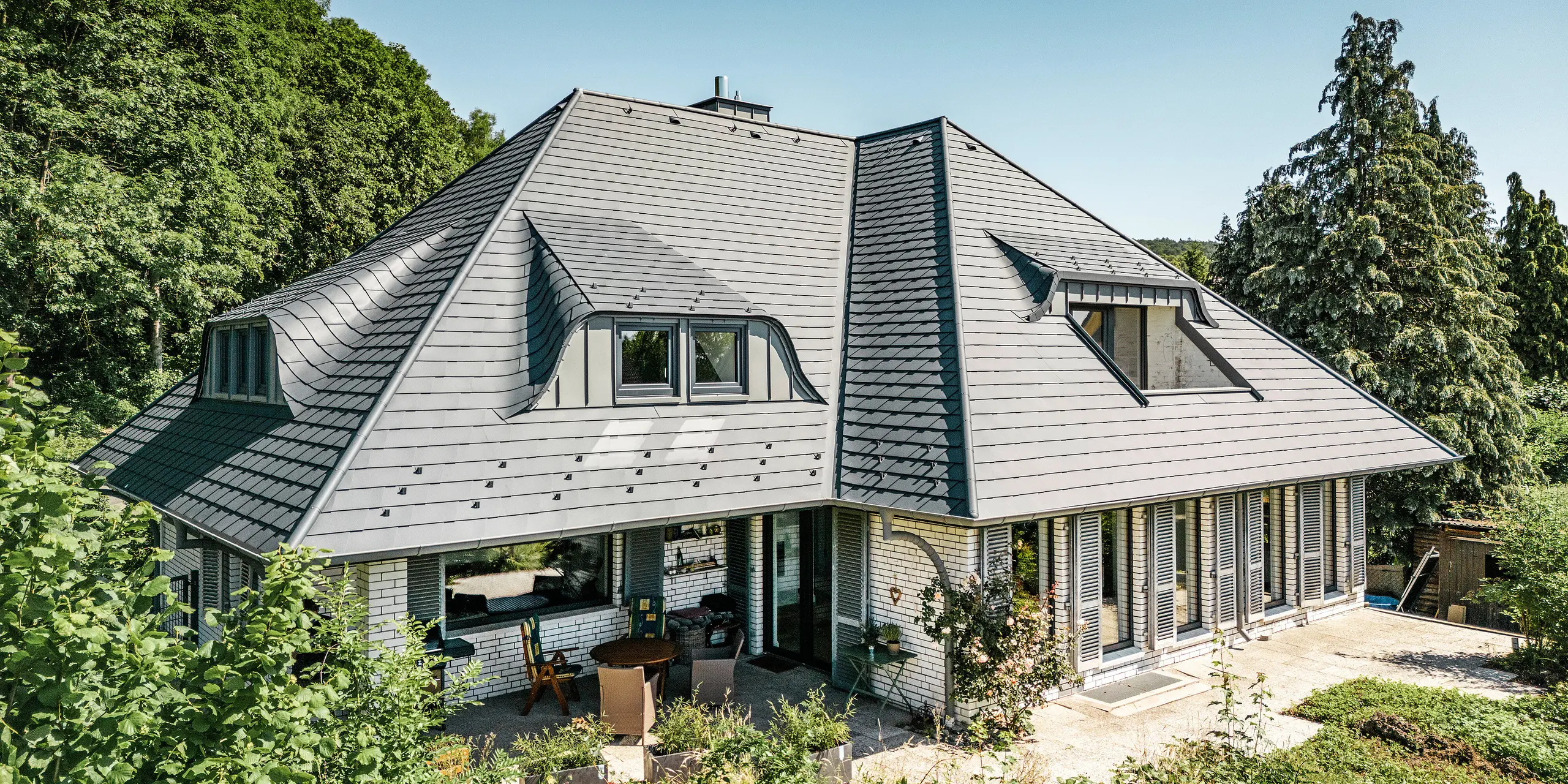 Moderne enfamiliehus i Einbeck, Tyskland, med PREFA tagsten i P.10 antracit. De holdbare PREFA tagsten i aluminium er kendt for deres kvalitet og holdbarhed. Tagkonstruktionen på bygningen understreger den individuelle arkitektur med gavlspidser, som fremhæves af PREFA tagstenes fleksibilitet. Tagafvanding i form af PREFA afløbsrør og tagrender er også holdt i antracit. Rundt om enfamiliehuse er der en stor og velplejet have samt en terrasse. Bag huset ligger en skov, der sammen med den blå himmel skaber en naturlig atmosfære.