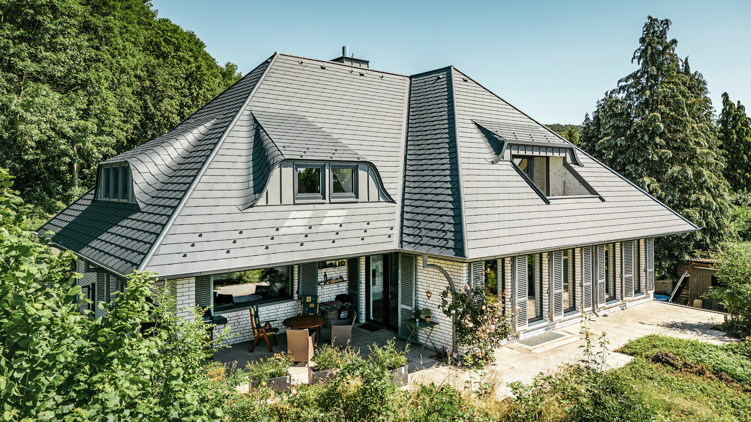 Moderne enfamiliehus i Einbeck, Tyskland, med PREFA tagsten i P.10 antracit. De holdbare PREFA tagsten i aluminium er kendt for deres kvalitet og holdbarhed. Tagkonstruktionen på bygningen understreger den individuelle arkitektur med gavlspidser, som fremhæves af PREFA tagstenes fleksibilitet. Tagafvanding i form af PREFA afløbsrør og tagrender er også holdt i antracit. Rundt om enfamiliehuse er der en stor og velplejet have samt en terrasse. Bag huset ligger en skov, der sammen med den blå himmel skaber en naturlig atmosfære.
