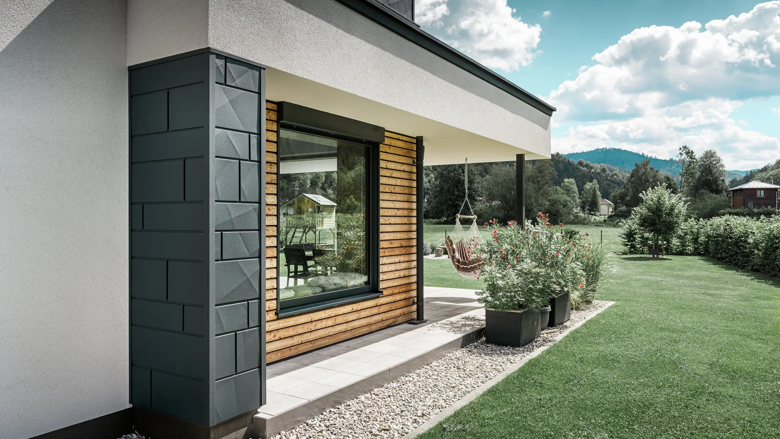Delvist billede af et nybygget parcelhus. Her er der brugt forskellige materialer på facaden. Den hvidpudsede facade fremhæves med PREFA aluminium Siding.X i antracit og vandrette træpaneler, som omgiver et stort vindue. På terrassen hænger en hængestol.