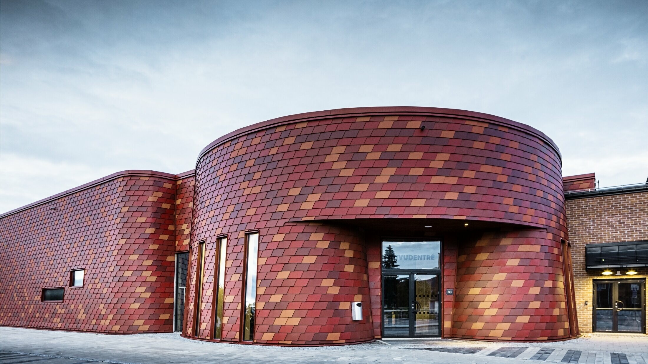Skøjtehal med fladt tag og svungen facade; facaden er beklædt med vægspån af aluminium fra PREFA i forskellige røde toner – oxidrød, teglrød, rødbrun, brunrød