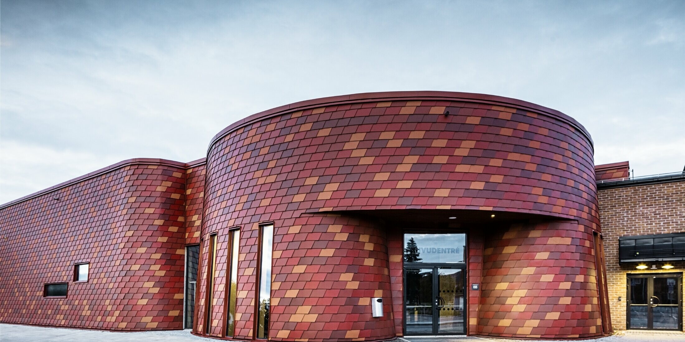 Skøjtehal med fladt tag og svungen facade; facaden er beklædt med vægspån af aluminium fra PREFA i forskellige røde toner – oxidrød, teglrød, rødbrun, brunrød